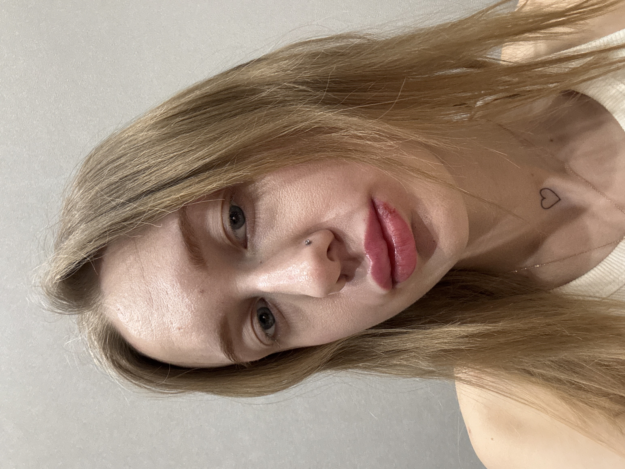 A close-up photographic image depicts a young adult female in Rostov-on-Don, Russia, looking directly towards the camera. The subject has long, straight, blonde hair and fair skin. Her eyes appear blue or grey, with a slight redness visible beneath the right eye. A small silver stud piercing is present on the right nostril. Her lips are full and tinted pink, with a visible lesion, exhibiting dried blood or scabbing, on the left side of the lower lip. A faint black outline of a heart tattoo is visible on the lower left side of her neck, above a thin gold-colored chain necklace. She wears a light-colored, possibly white or off-white, ribbed top. The background consists of a plain, light grey or off-white wall with a subtle textured finish. The overall scene appears to be an interior setting, likely a room.