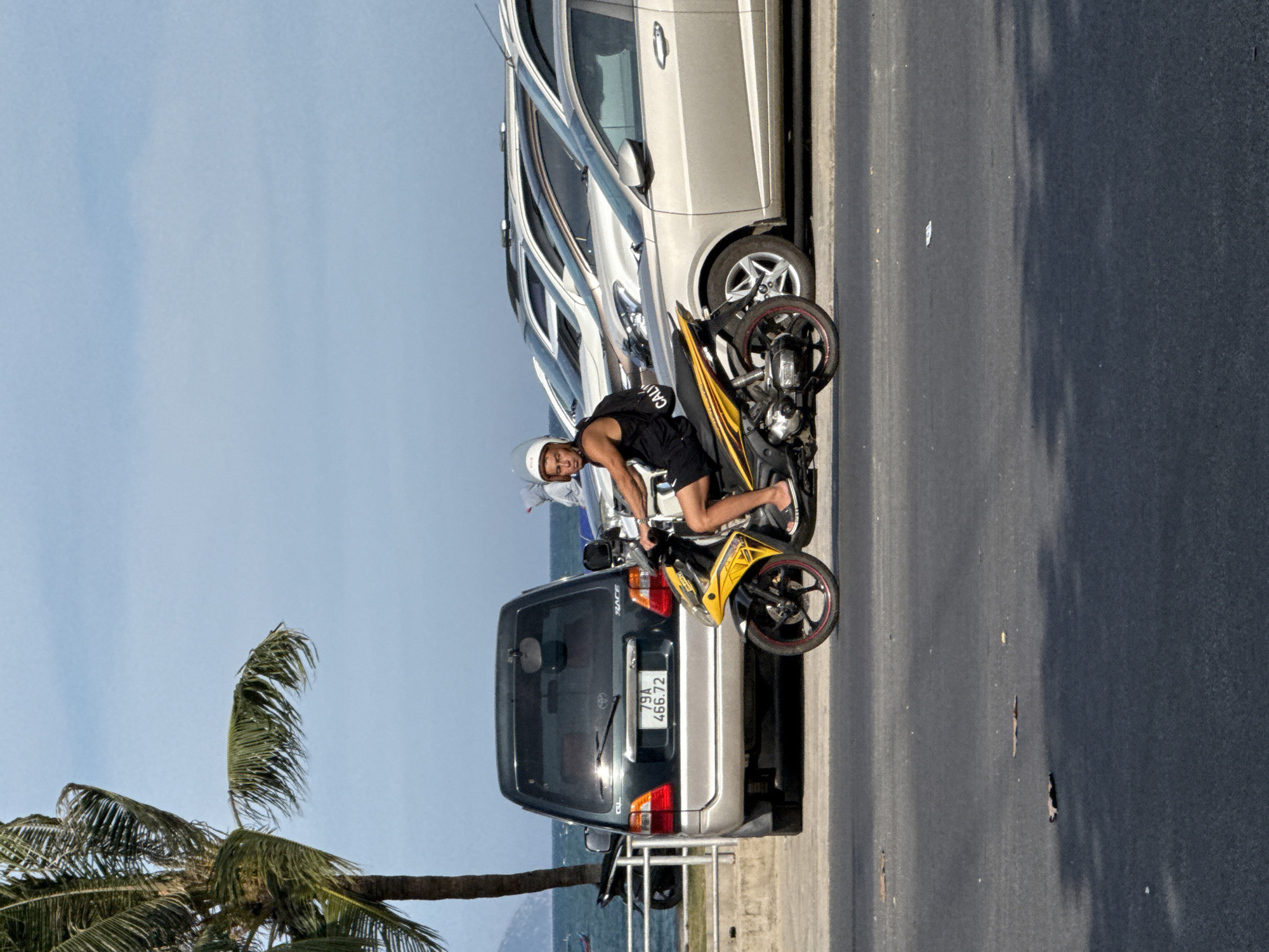 A young man wearing a white helmet and black tank top rides a yellow and black motorcycle on a street in Bac Nha Trang Ward, Vietnam. He is looking towards the right. The motorcycle has a black backpack strapped to its back. The man is wearing flip-flops. In the background, there are several cars parked along the street. A dark-colored van with a license plate reading "79A 466.72" is parked to the left of the motorcycle. A palm tree is visible on the far left of the image. The sky is clear and blue, and the sea is visible in the distance. The street is paved and appears to be in good condition.