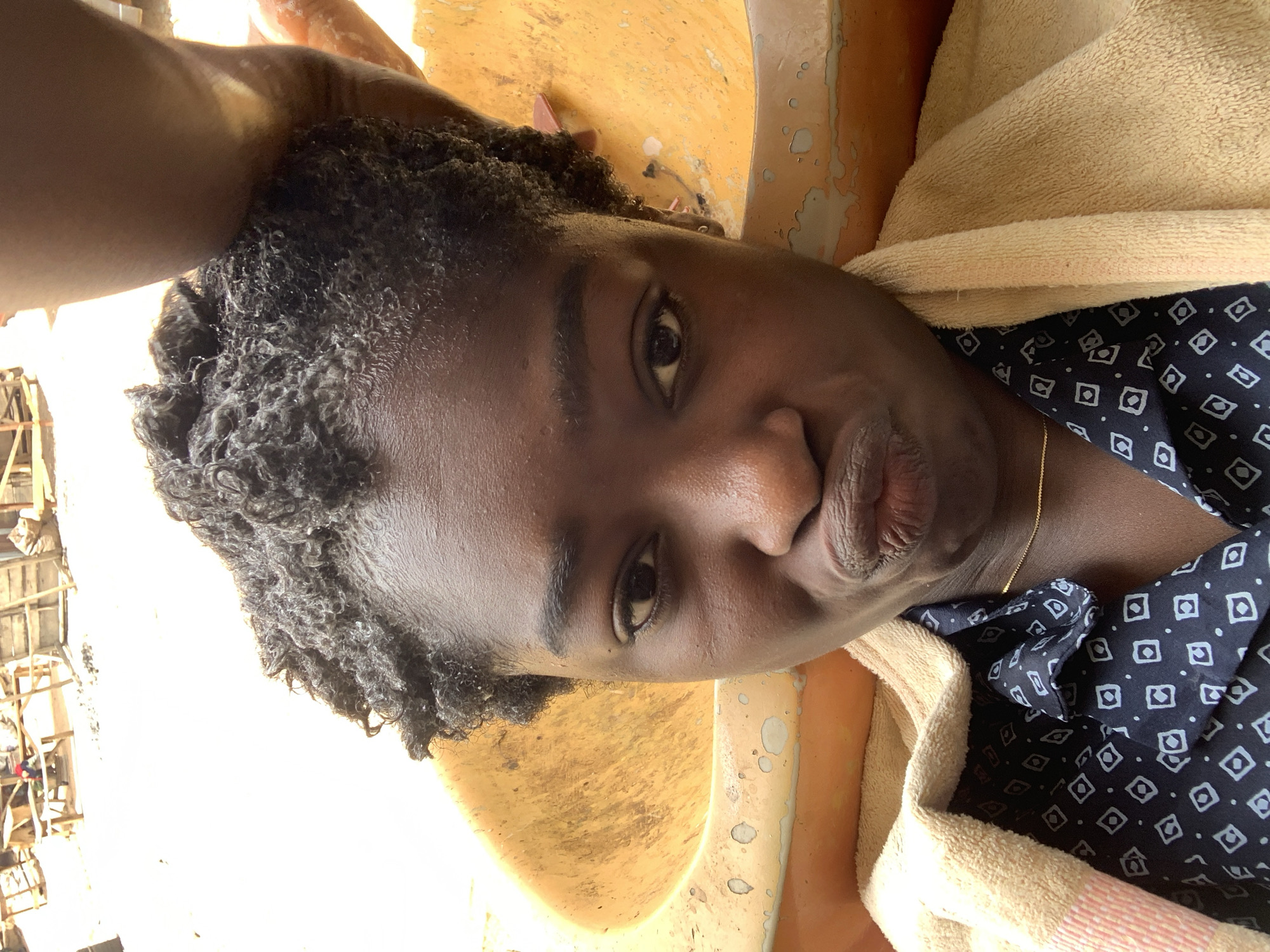 The image shows a dark-skinned person with short, dark, damp, curly hair, possibly in a salon or washroom. They are wearing a dark blue shirt with a geometric pattern and have a light-colored towel draped around their shoulders. The person is in what appears to be a sink or basin. The location appears to be Rafin Par, Nigeria, as specified, but there is no direct evidence of the location within the image. Outside the window, there appears to be scaffolding or wooden construction.