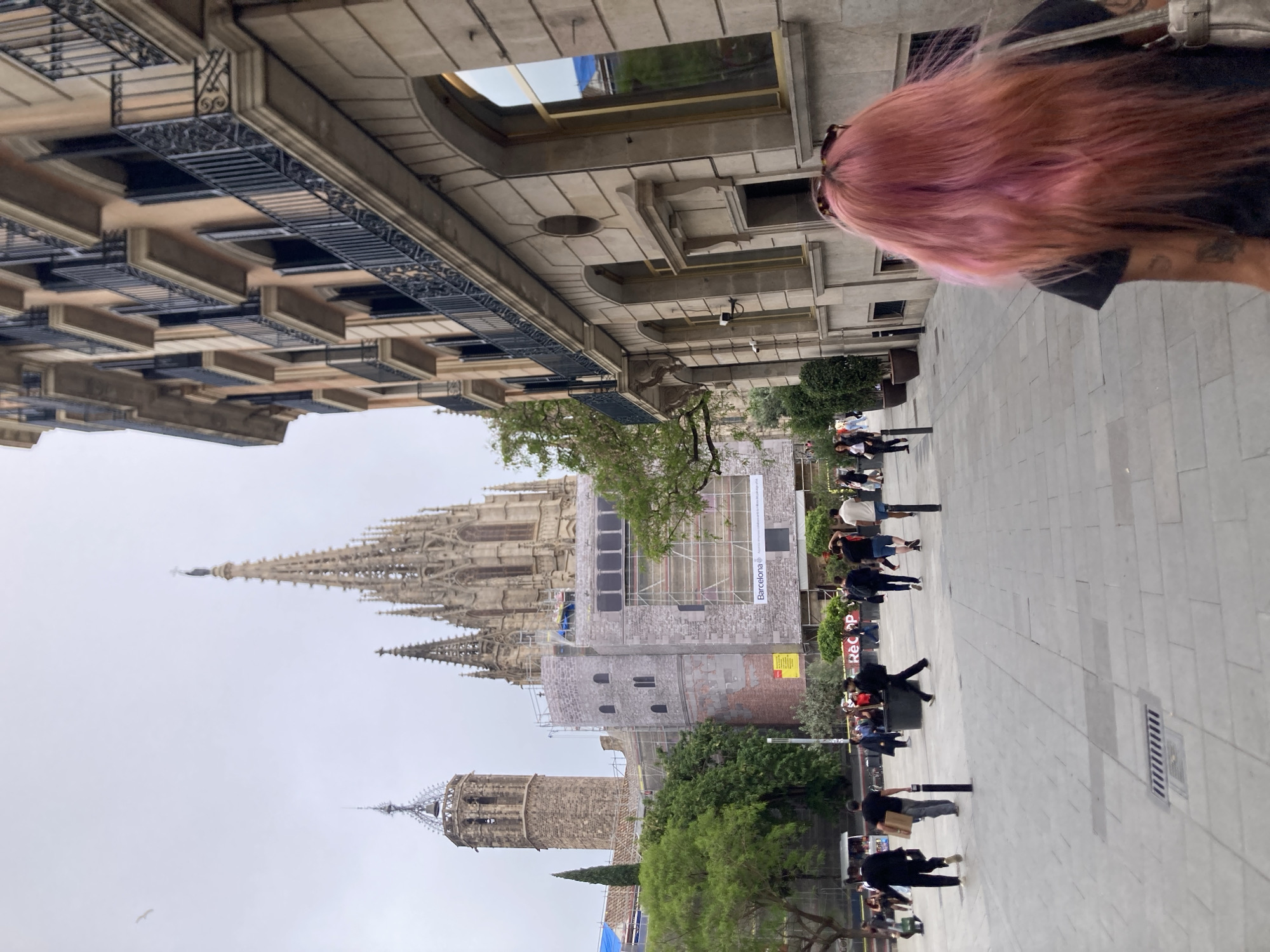 The image shows an outdoor scene in Barcelona, Spain. A woman with pink hair is in the foreground on the right. A street stretches away from her, with pedestrians walking along it. On the left, tall buildings line the street. One building is under construction, surrounded by scaffolding. In the distance, the spires of a large, ornate cathedral can be seen against a gray sky. The overall impression is a typical urban scene in a historical city.