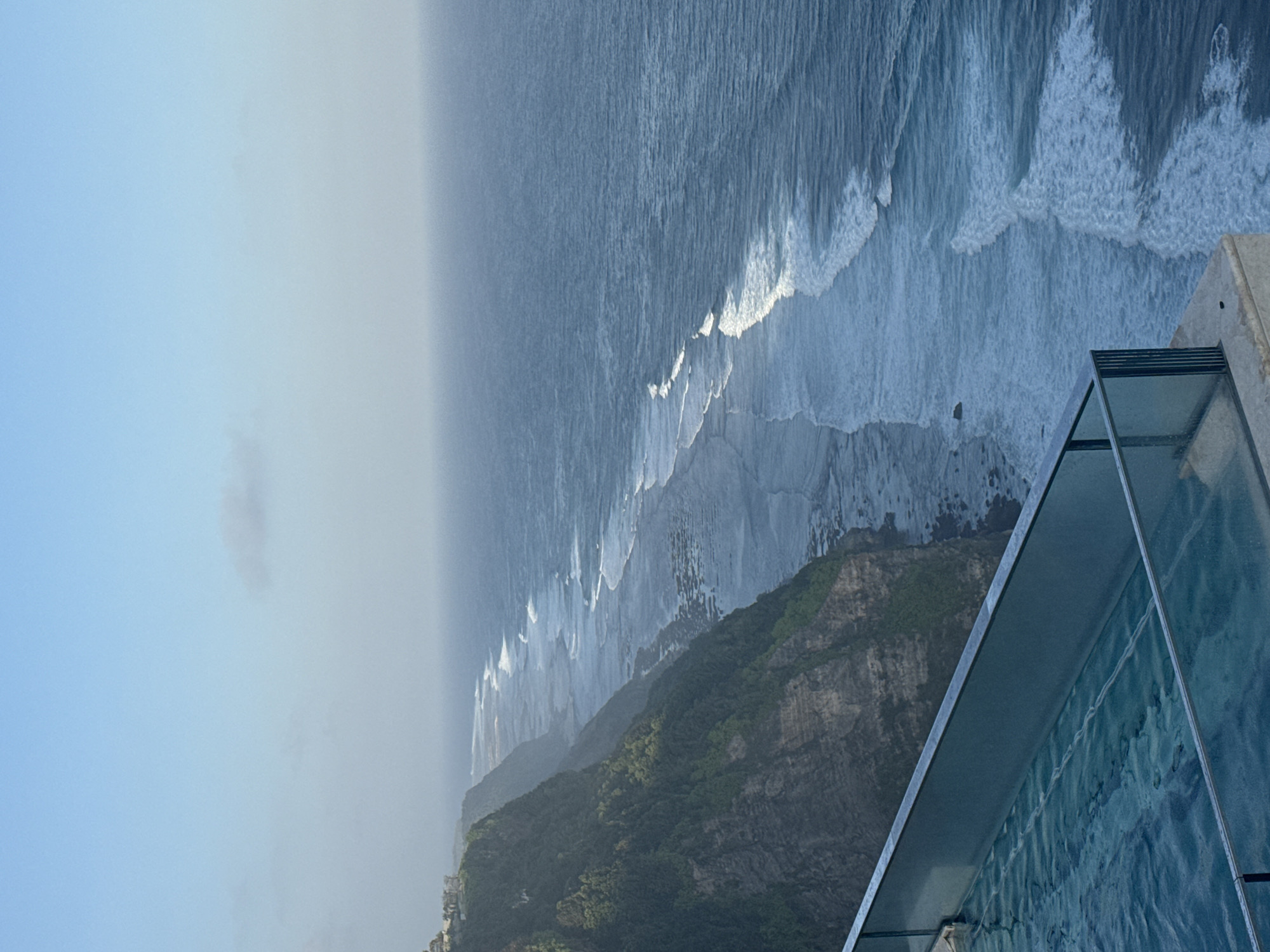 An infinity pool with a glass railing overlooks a dramatic cliffside with dense green foliage meeting the choppy ocean below. The ocean stretches to the horizon, with white waves crashing along the rocky shore. The sky above is a pale, hazy blue. A few buildings are visible on the cliff top in the distance.