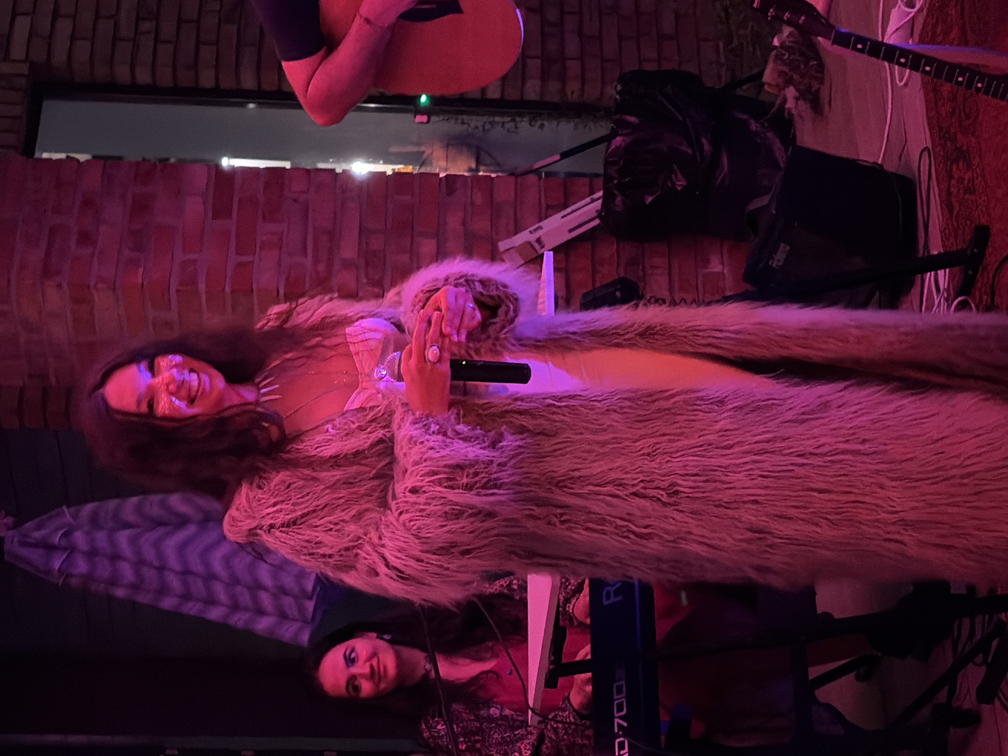 In a dimly lit outdoor setting, possibly a backyard or patio, illuminated by vibrant pink lighting, a woman stands center frame, holding a microphone and wearing a long, shaggy, light-colored faux fur coat over a light-colored dress. She has dark, long, wavy hair and is smiling. To her left, another woman with dark hair sits at a keyboard, wearing a patterned top and looking towards the main subject. To the right of the main subject, a person is partially visible, playing an acoustic guitar. In the background, a brick wall, a closed umbrella, a black trash bag, and a speaker are visible. The overall scene suggests a musical performance or gathering. The location is specified as Shebanovo, Russia.