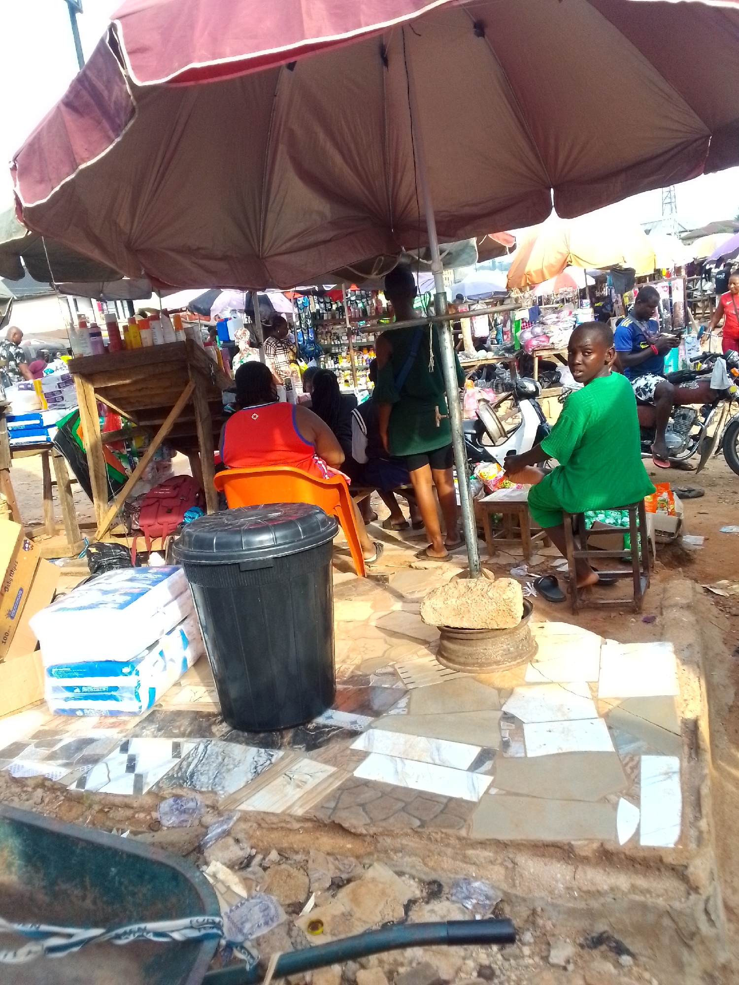 The image depicts a bustling street scene, likely a market area, in Orlu, Nigeria. The foreground shows a partially visible wheelbarrow with a dark-colored handle. Above that, a rough, tiled pavement leads to a dark, plastic trash bin sitting next to stacks of boxed goods, possibly diapers or wipes. A large, brown market umbrella dominates the upper part of the frame, providing shade to several vendors and customers. People are gathered around tables displaying various goods. A young boy in a green outfit sits on a small stool, looking toward the camera. Other people are seated on chairs nearby, possibly engaged in conversations or transactions. In the background, there are more stalls and individuals, and at least one motorcycle is visible. The overall atmosphere appears to be lively and crowded, typical of a busy market environment.