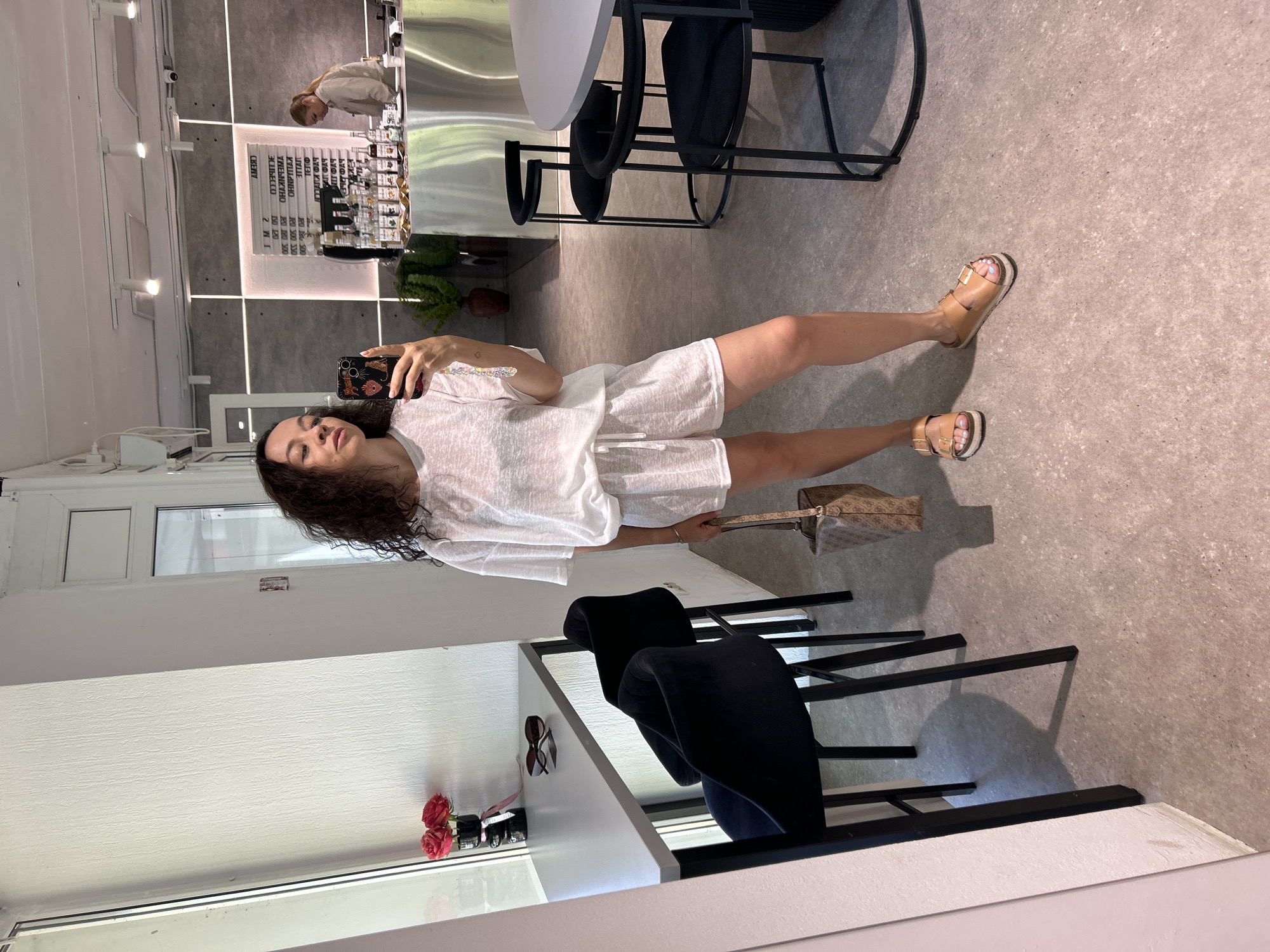In a cafe setting, a woman stands by a window, taking a mirror selfie. She is wearing a white t-shirt and shorts set, brown sandals, and is holding a monogrammed brown bag. To her left, a narrow table is set against the window, with two black bar stools. On the table, there are two red roses in a vase and a pair of sunglasses. In the background, a coffee bar is visible with bottles of syrup and a menu board. A second woman, wearing a beige jacket, stands behind the counter. The cafe appears to be located in Chaikovsky, Russia. The lighting suggests daytime.