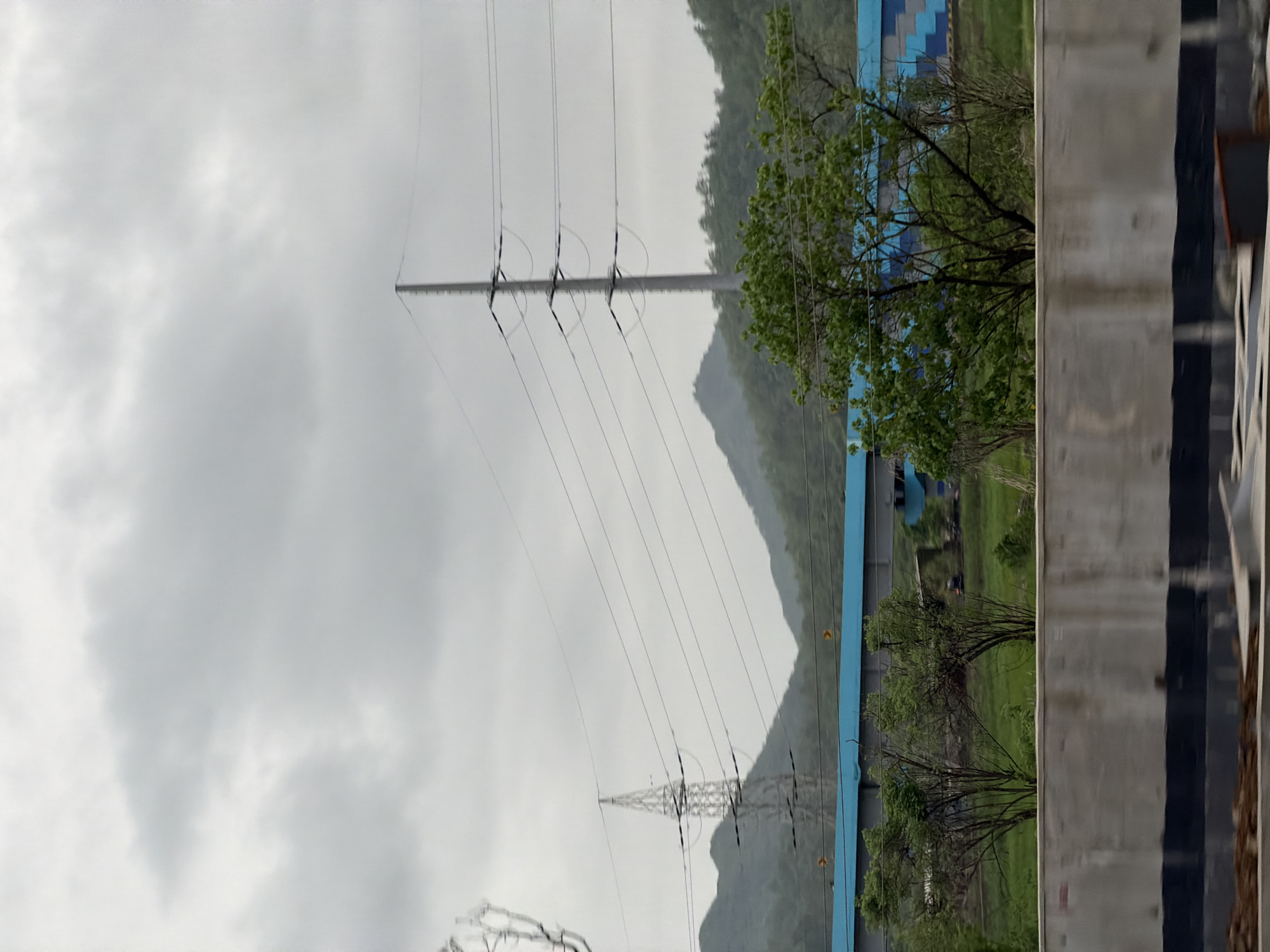 The image depicts a cloudy day in Saphale, India. In the foreground, a concrete barrier with black stripes runs horizontally across the frame. Behind the barrier, several trees with green foliage are visible. A long, blue and grey structure stretches across the mid-ground, with a bridge element visible within it. In the background, rolling green hills are partially obscured by a layer of mist or clouds. Power lines crisscross the scene, supported by tall, metal pylons. One pylon is a lattice tower, while another is a single, tall pole. The overall atmosphere is overcast and somewhat muted. No people or animals are visible in the scene.
