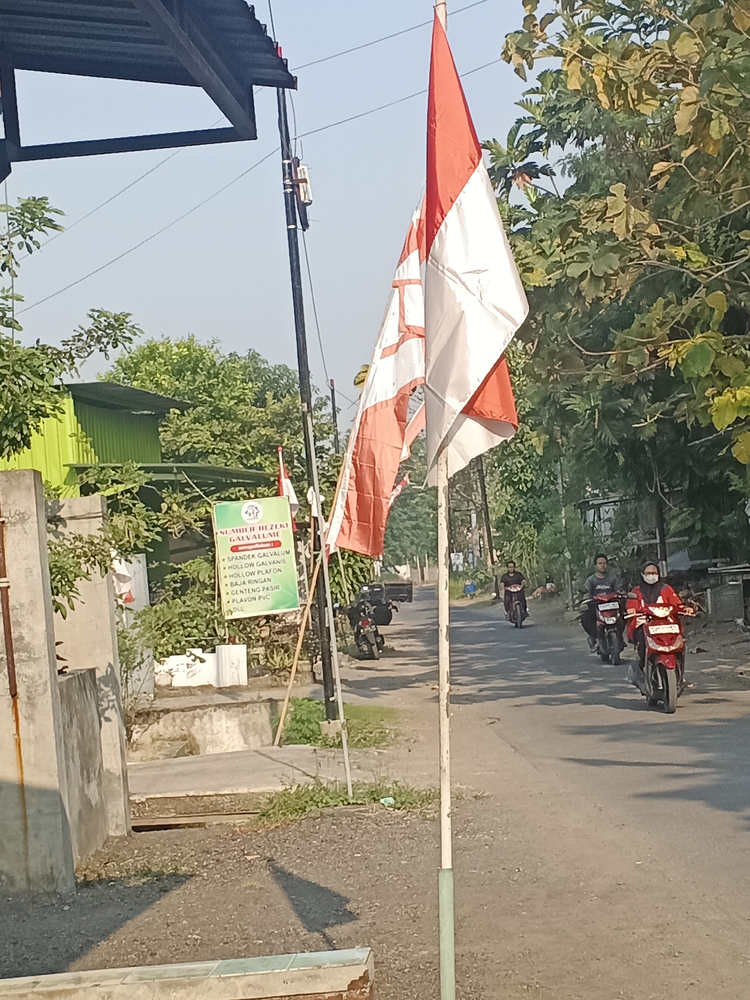 This image depicts a street scene in Krian, Indonesia, dominated by the Indonesian flag, which is red and white, flying prominently. A sign is visible on the left, advertising "SUMBER REZEKI GALVALUM" and listing various building materials such as "SPANJEK GALVALUM," "HOLLOW GALVANIS," "HOLLOW PLAFON," "BAJA RINGAN," and "GENTENG PASIR." The street is paved and appears to be a local road. Several individuals on motorcycles are traveling down the street. One person in the foreground on a red motorcycle is wearing a mask and a red top. Another person on a motorcycle behind them is wearing a grey shirt. Further down the road, another individual on a motorcycle is visible. The environment is characterized by lush green trees lining the street and a bright, clear sky, suggesting daylight. Buildings with green metal siding and concrete structures are also present in the background.