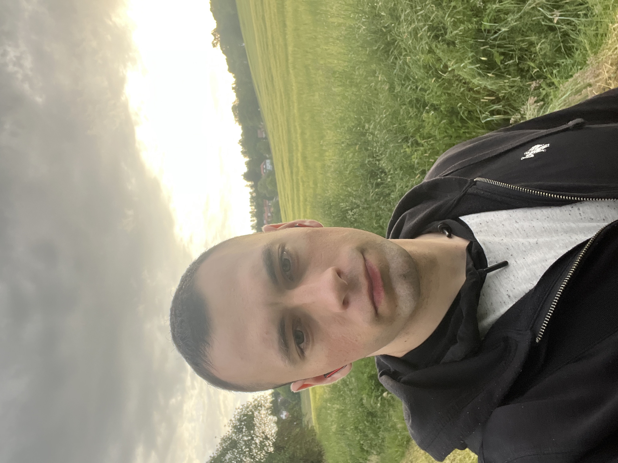 A man is taking a selfie. He is wearing a black zip-up hoodie with a white shirt underneath. He has short, dark hair and light skin. Behind him is a field of green grass and trees, under a sky with a mix of clouds and sunlight.