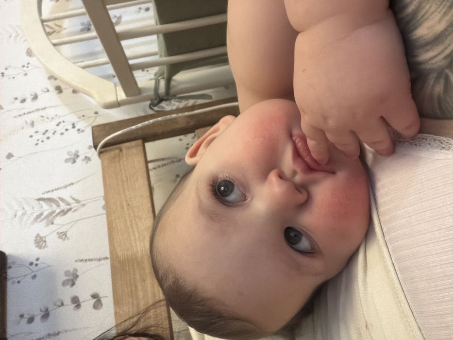 A close-up shot reveals a baby lying on their side, possibly in a crib or on a bed with floral-patterned sheets. The baby's face is the focal point, with rosy cheeks and bright eyes looking upwards. Their tiny fingers are near their mouth, suggesting they might be teething or exploring their hands. A glimpse of the baby's arm and a white garment are visible, adding context to the scene. A wooden crib rail is visible in the background.