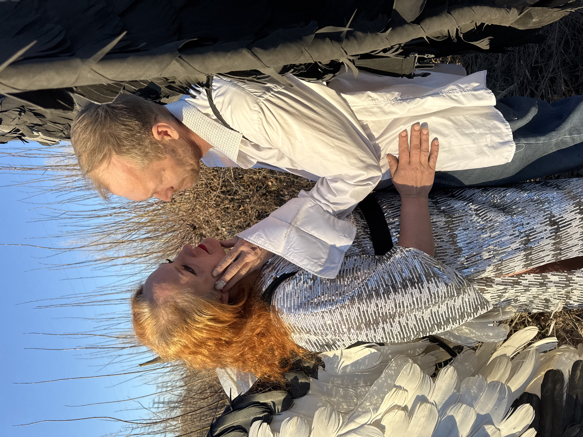 A man and a woman are in close proximity to each other, lying on the ground in an outdoor setting. The man is wearing a white shirt and blue jeans. The woman has red hair and is wearing a silver sequined top and a black undershirt. The woman is touching the man's face with her right hand. The couple is lying on what appears to be grass or vegetation with some visible bare branches behind them. Large black and white feathered objects, possibly wings or part of a costume, are positioned around them. The scene is set against a clear blue sky. The location is Chaikovsky, Russia.