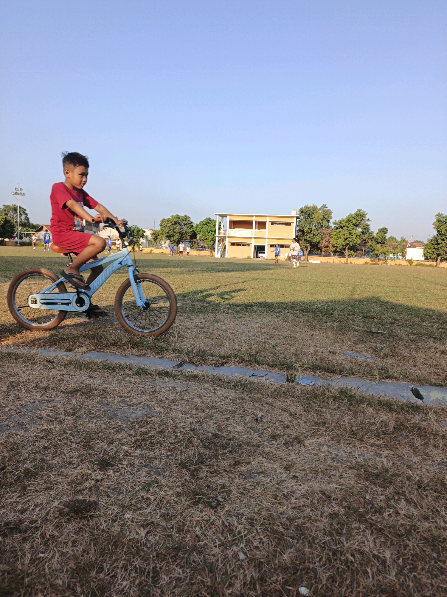 A young boy, appearing to be around 8-10 years old, rides a light blue bicycle on a grassy field. He is wearing a red t-shirt and red shorts, with sandals on his feet. The bicycle has brown-rimmed wheels and appears to be a BMX-style bike. In the background, there is a yellow and white school building with trees surrounding it. Several other children, some in blue and white uniforms, are visible on the field, possibly playing a sport. The sky is clear and blue, and the lighting suggests it is late afternoon. The ground cover is a mixture of dry, straw-like grass and some greener patches. The scene appears to be a schoolyard or sports field in Mojokerto, Indonesia.