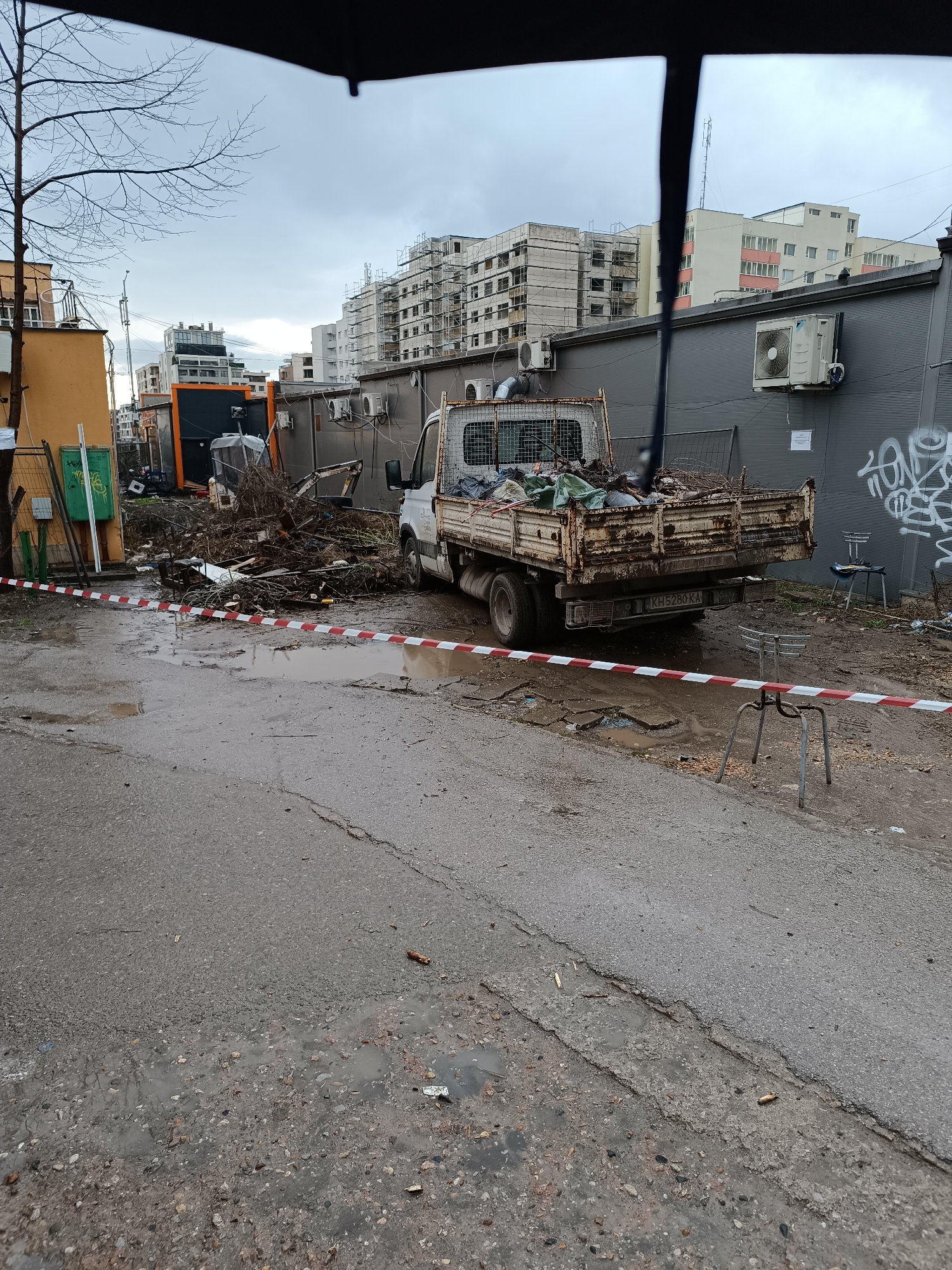 The image depicts a street scene in Sofia, Bulgaria, likely a back alley or less-maintained area. The street is wet with puddles, and the pavement is cracked and broken. A small, weathered white truck with a flatbed filled with branches, leaves, and general debris is parked on the right side of the image. The license plate number on the truck is KHS280 KA. A pile of similar debris is present in front of the truck, partially blocked off by red and white striped caution tape. Two metal stools are near the caution tape. Behind the truck is a grey wall with graffiti and air conditioning units attached. Further back, there are several buildings, including one under construction with scaffolding. To the left, there is an orange building and a small construction vehicle. The sky is overcast and grey. The overall impression is one of urban decay and possible construction or cleanup activity.