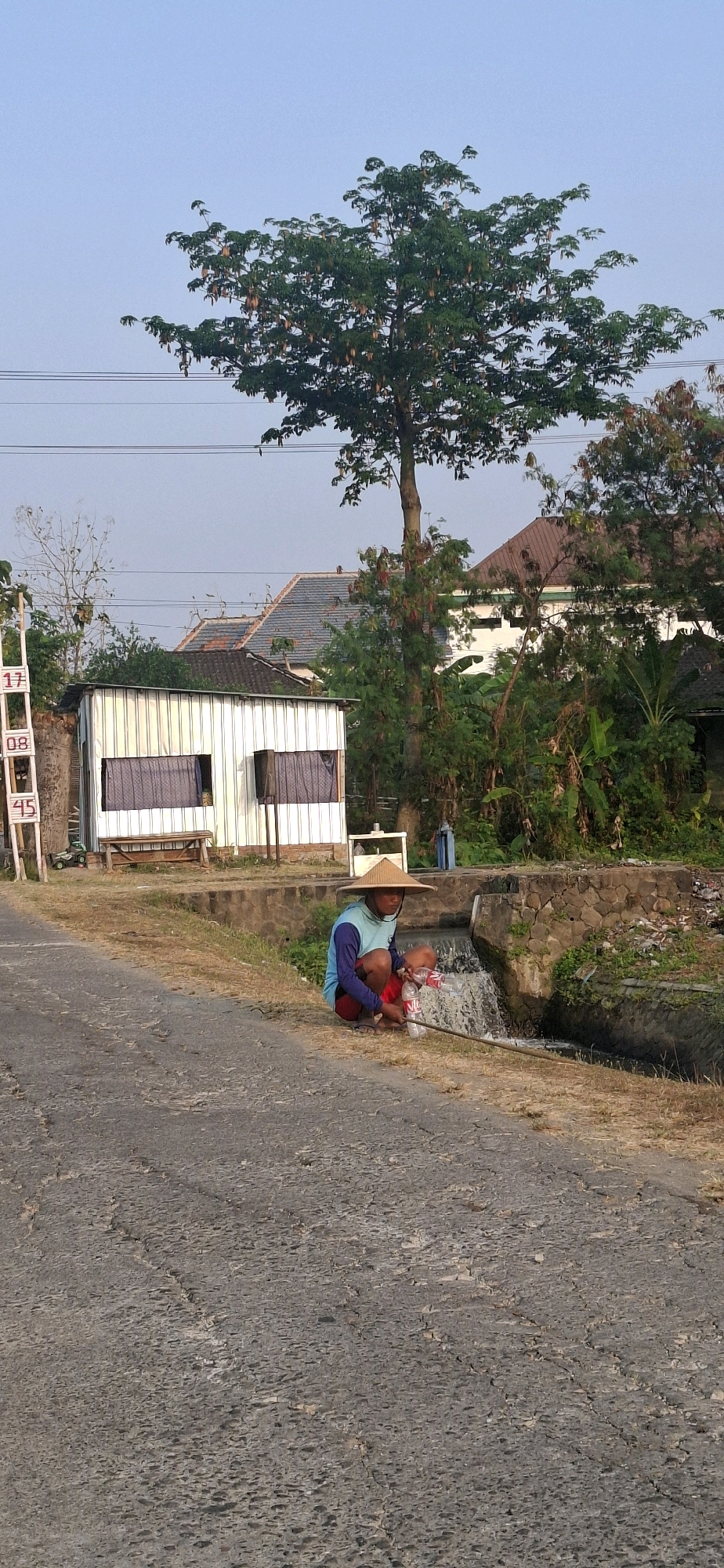 In Desa Ngesrep, Indonesia, a person wearing a conical hat and a blue and red outfit is crouched near a small water channel, holding a fishing rod and two plastic bottles. The person appears to be fishing or collecting something from the water. Behind them, a simple white corrugated metal building with two windows covered by purple curtains stands beside a road. A ladder with numbers "17," "08," and "45" is visible to the left of the building. The scene is surrounded by greenery, including trees and bushes, and residential buildings with tiled roofs are visible in the background. The sky is clear and light blue, suggesting it is daytime.