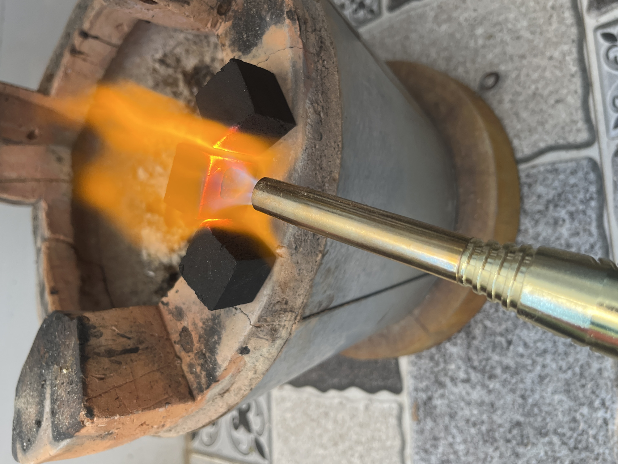 The image shows a close-up of a cooking setup in Bac Nha Trang Ward, Vietnam. A traditional Vietnamese stove, made of clay and metal, is being used to burn what appears to be charcoal briquettes. A metallic torch is directed at the briquettes, igniting them and producing a vibrant orange flame. The stove rests on a tiled surface. No people are visible in the frame.
