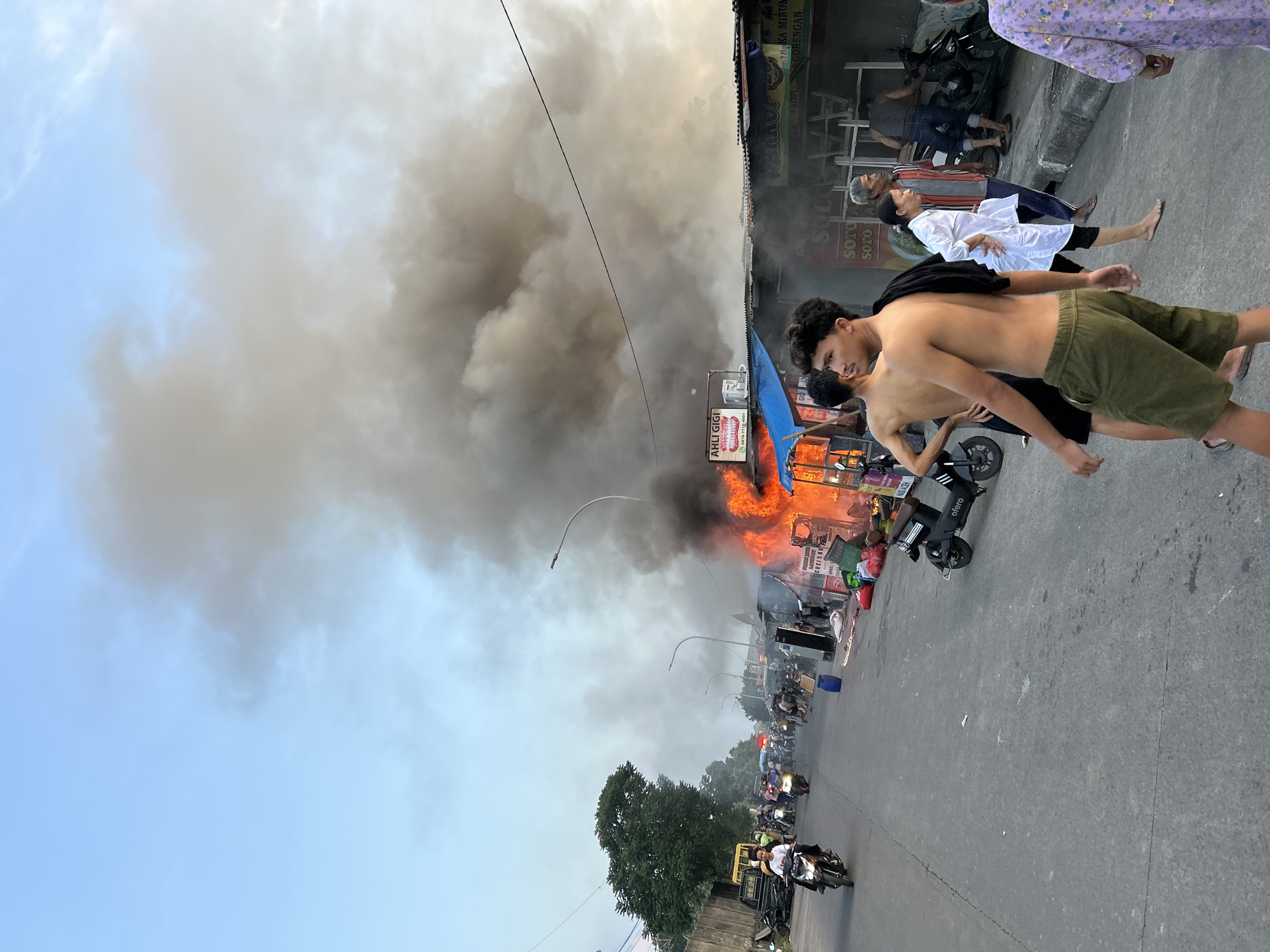 In an urban street scene, a significant fire engulfs several market stalls, producing a large plume of dark smoke that billows into the sky. The fire is visible on the right side of the image, characterized by bright orange flames and illuminated by the twilight sky. In the foreground, a young shirtless man wearing green shorts walks away from the camera, his back to the viewer. To his left, another shirtless young man in black shorts walks in the same direction. Behind them, a woman in a white top and dark pants walks towards the fire, looking concerned. Further back, several people observe the fire, some standing near motorcycles. The street is paved, and on the left side, there is a wall and trees. A line of motorcycles and people are visible further down the street, suggesting a busy marketplace. The stalls appear to be made of corrugated metal and wooden structures, some with visible signage, indicating commercial activity. Several motorbikes and some blue plastic barrels are scattered around the scene. The overall atmosphere is one of chaos and concern due to the ongoing fire.