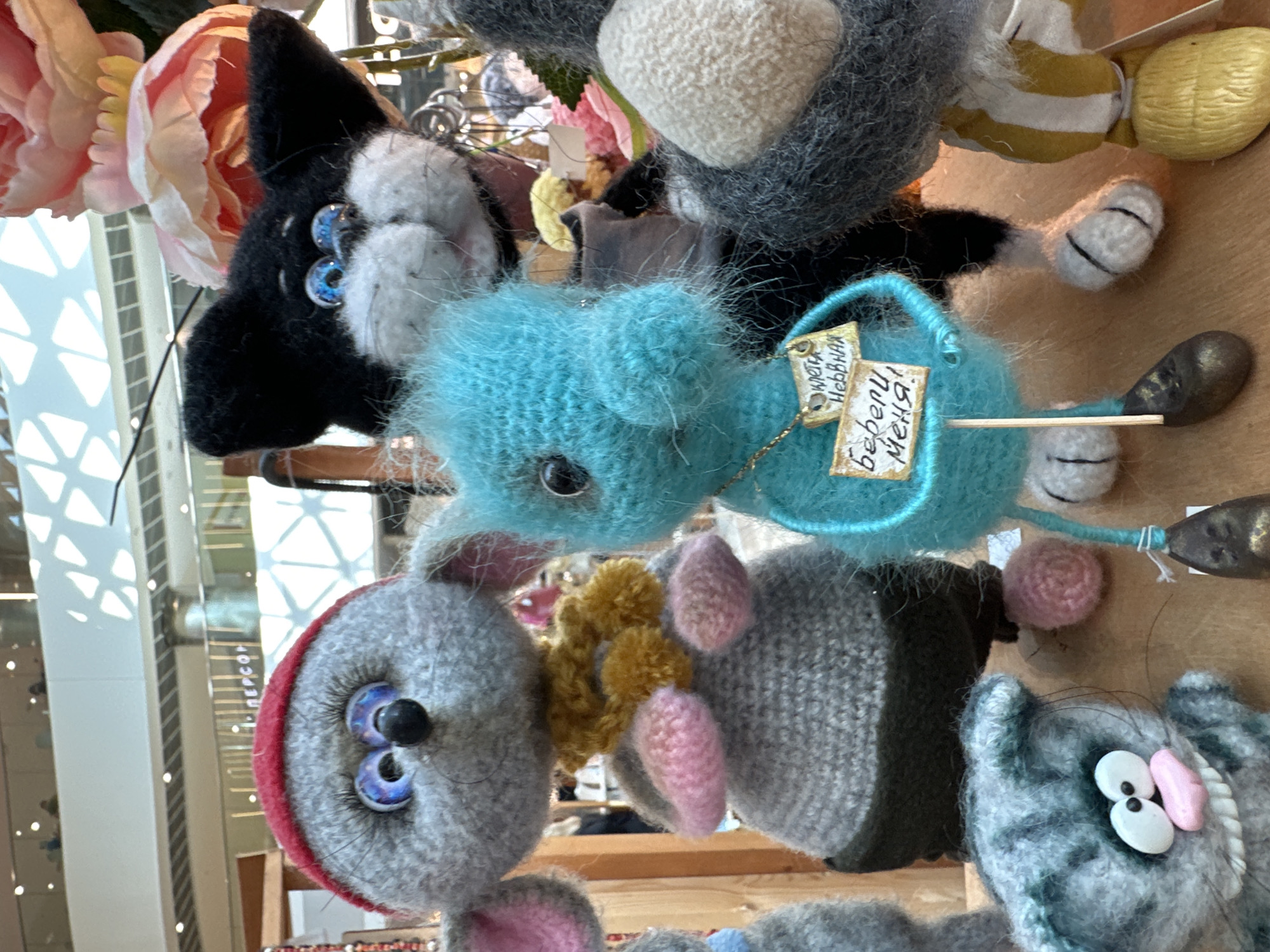 The image captures a collection of handmade plush toys, likely for sale, arranged on a wooden surface. The toys are whimsical and detailed, with a variety of characters including a grey mouse wearing a red hat and yellow scarf, a black and white cat with blue eyes, a turquoise creature with a sign around its neck, and a grey striped cat with a wide grin in the foreground. In the background, there is a blurred view of an indoor environment, suggesting a shopping mall or a market with multiple levels and architectural features. While the scene is detailed and visually engaging, there is no specific information within the image that identifies it as being located in Sochi, Russia. The presence of Russian text on the turquoise toy's sign, "Цветет Нежная", and "Весели Меня!", suggests a Russian origin for the toys or the location where they are being sold.