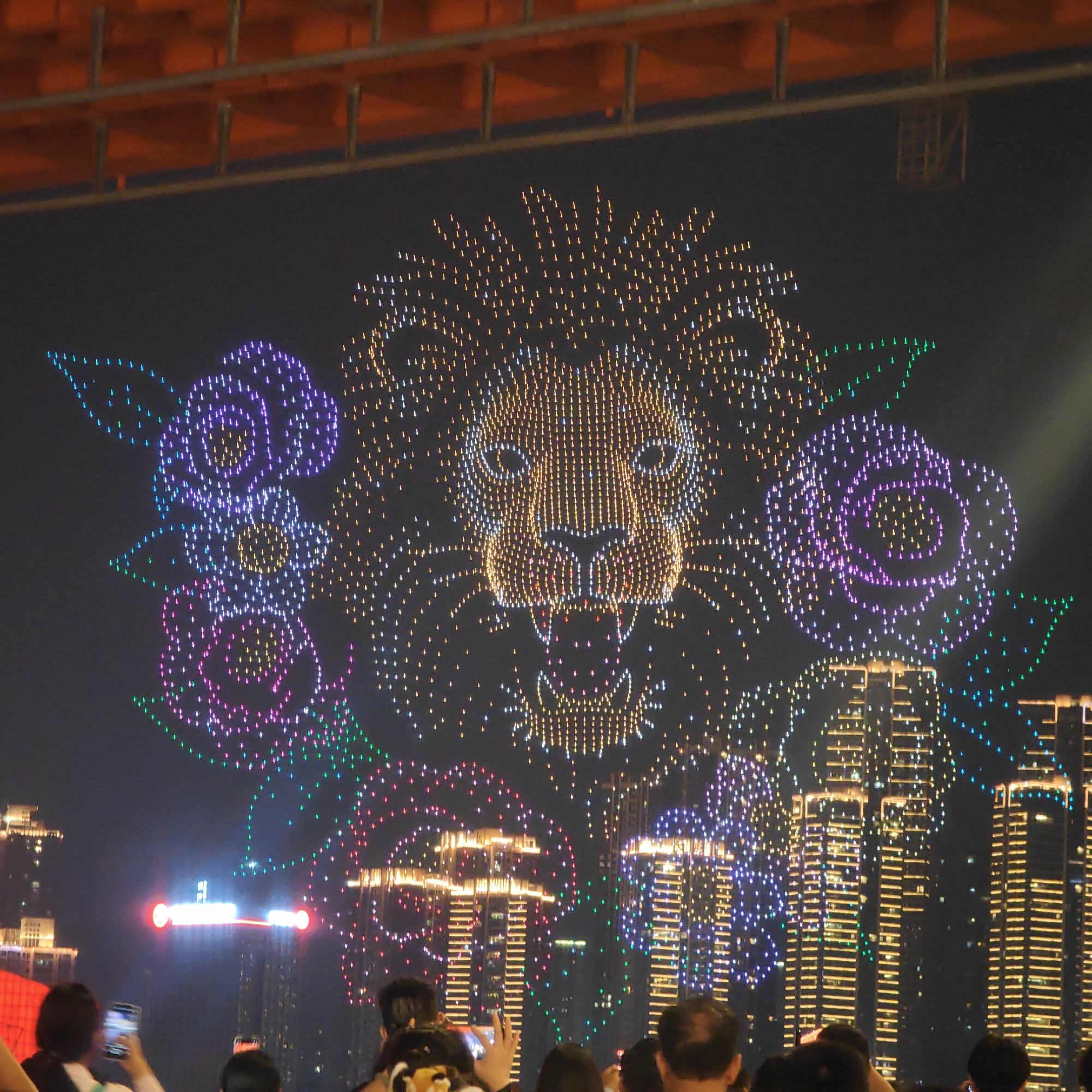 At night in the Yuzhong District, China, a large light display is visible, depicting a lion’s head surrounded by flowers. The display is set against the backdrop of tall buildings, some of which are illuminated. A crowd of people is present, some holding up their phones to capture the spectacle. An orange structure is visible in the upper part of the frame.