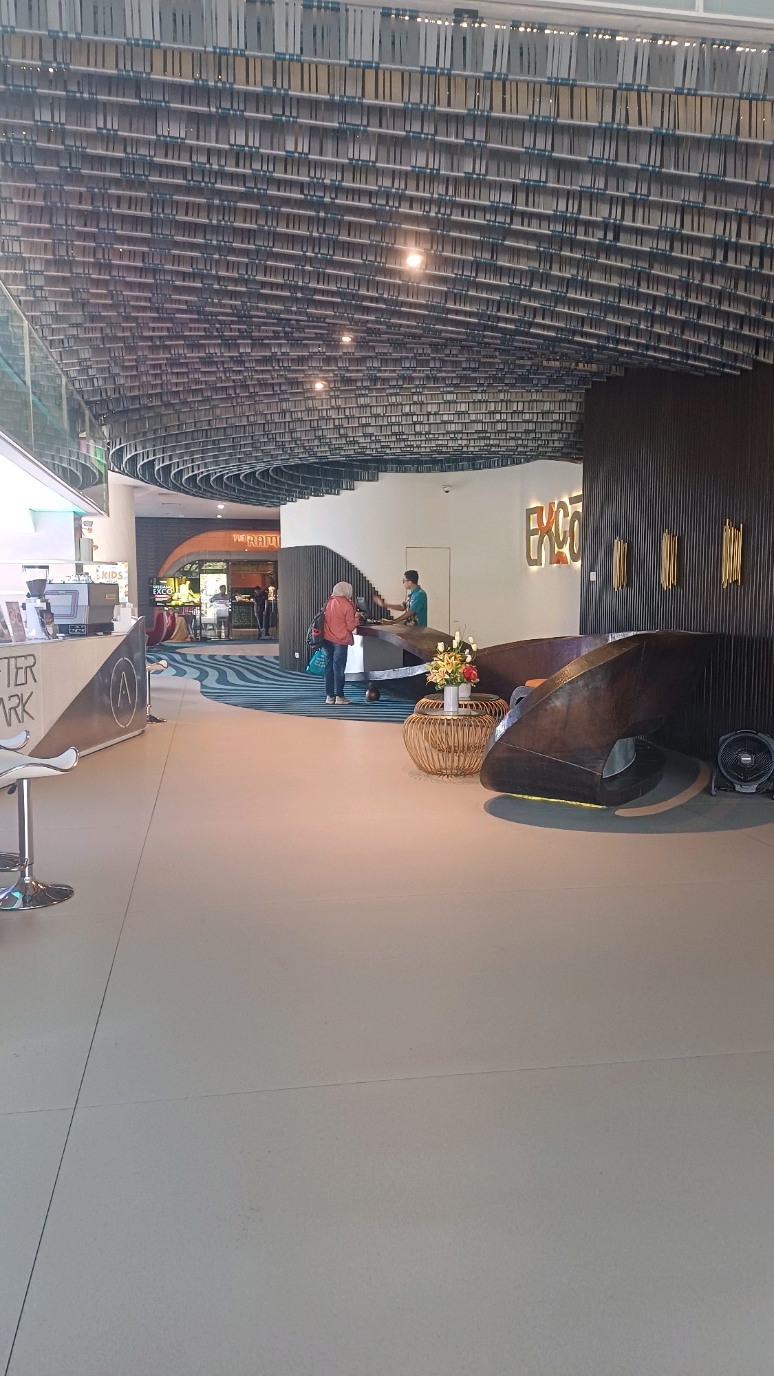 The image depicts the lobby of a hotel or establishment with modern, sculptural design. The ceiling features an intricate, layered structure of metal elements, creating a visually complex canopy. A reception desk, with an organic, wave-like form made of dark wood or metal, is centrally located. Two individuals are visible near the reception desk; one appears to be a staff member, wearing a green shirt and standing behind the desk, while the other, a patron in a red jacket, stands in front of it. In the foreground, a light-colored floor with visible grout lines extends towards the viewer. To the left, a counter with a coffee machine and a stool suggests a cafe or waiting area, with text that includes "AFTER PARK." Further in the background, a staircase is visible, and behind it, a sign reads "THE RAMI." The overall ambiance is contemporary and stylish, with a neutral color palette accented by the warm tones of the reception desk and the signage. The scene is set indoors, in what appears to be a public space with a high ceiling.
