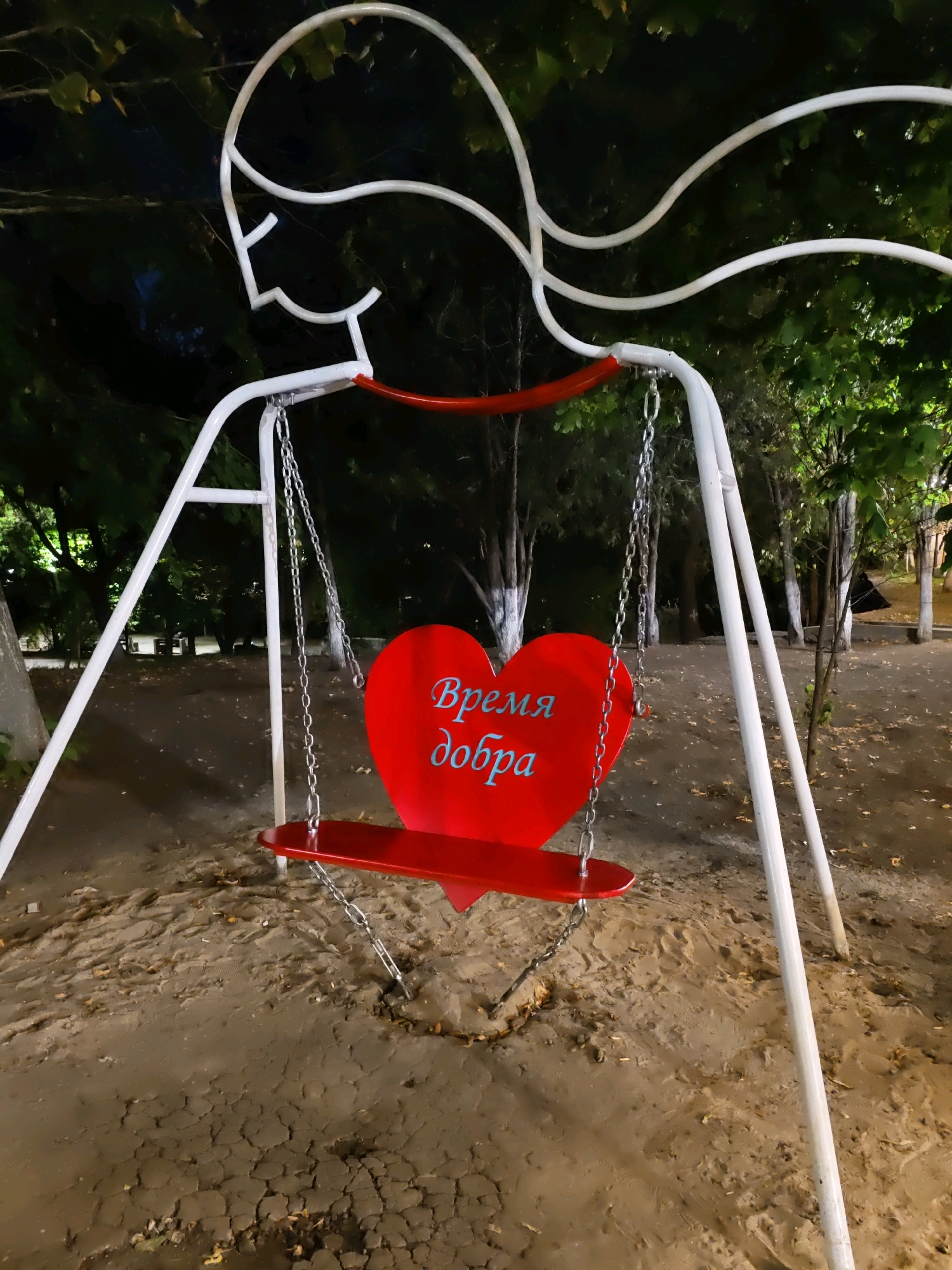 A red heart-shaped swing seat with blue text reads "Время добра" (Time for kindness) hangs from a white swing set with abstract human-like figures. The swing set is located outdoors in a park-like setting at night. The ground beneath the swing is sand and dirt, with some fallen leaves. Trees with dark foliage are visible in the background.