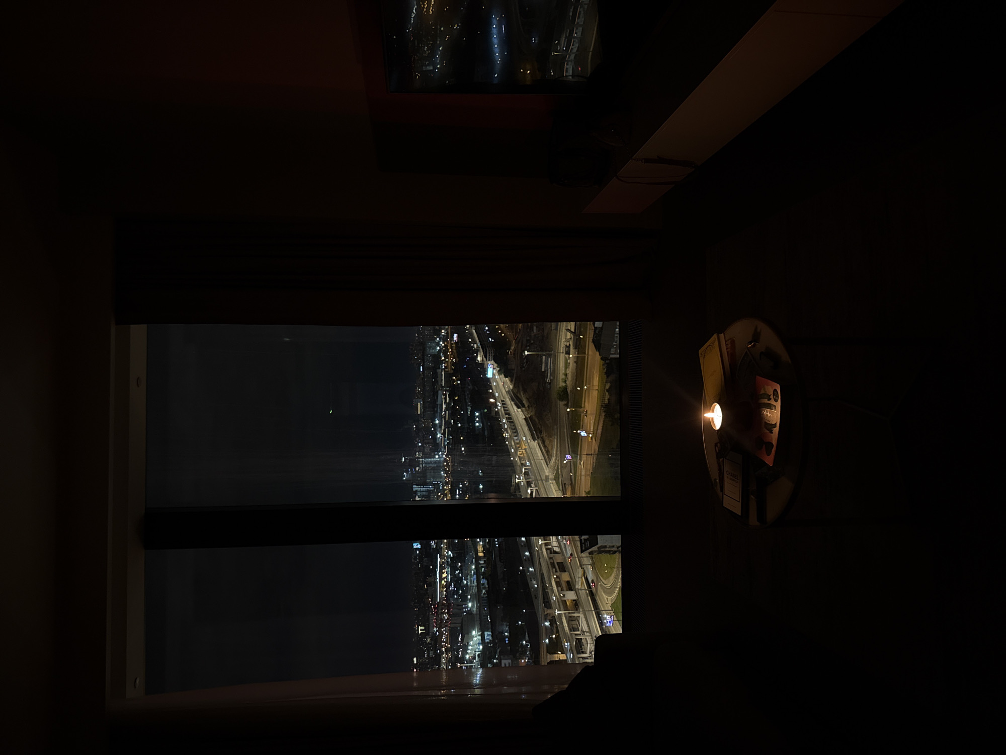 A dimly lit interior room overlooks a night cityscape, likely in Khimki, Russia, based on the visible urban infrastructure. The view is framed by a window with a horizontal divider, showing illuminated city streets, buildings, and highways. A round table is visible to the right, lit by a single candle and displaying magazines. The environment is dark with a focus on the illuminated cityscape.