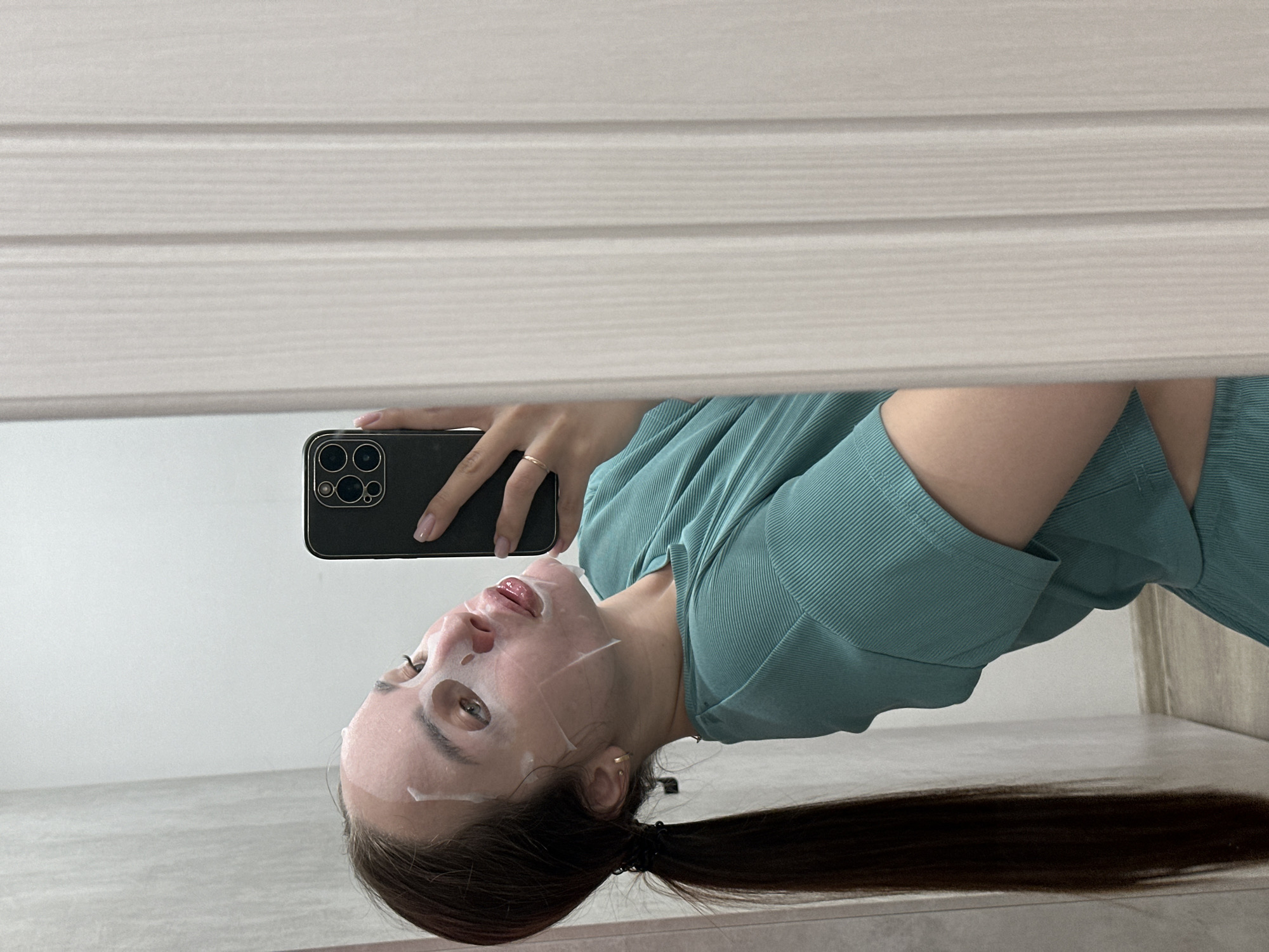 The image captures a mirror selfie of a young person, likely female, engaged in a self-care routine. The individual's face is covered with a white sheet face mask, featuring cutouts for the eyes, nose, and mouth. She is wearing a teal-colored, ribbed short-sleeved top. Her long, dark hair is pulled back, with a significant portion cascading downwards in the reflection.
In her left hand, which bears a gold ring on the ring finger, she holds a black smartphone with a prominent triple-camera system, presumably used to take the picture. Small gold stud earrings are visible in her ears.
The reflection in the mirror reveals an indoor setting. The background consists of a plain, light-colored wall, and a light-colored floor, possibly tiled or wood laminate, is visible beneath her. At what would be the top right if the image were upright, the reflection shows horizontal light-colored blinds or wooden paneling. The scene depicts a moment of personal care indoors. No discernible text is visible. The location context for this activity is Chaikovsky, Russia.