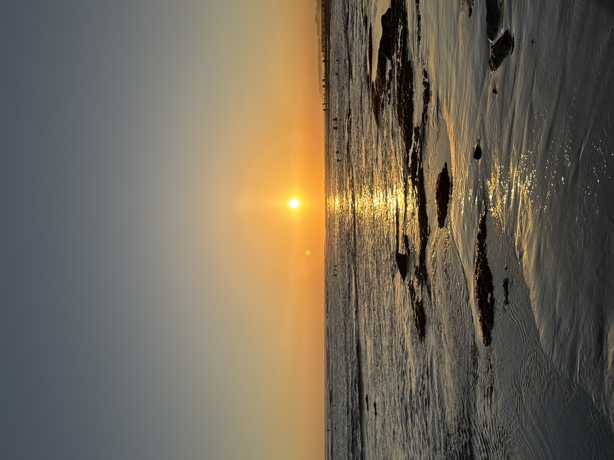 The image depicts a serene sunset over the ocean in North Kuta, Indonesia. The sun, a bright orb of orange, is positioned in the center of the frame, casting a shimmering golden path across the water. The sky transitions from a pale blue at the top to a warm orange glow near the horizon. Gentle waves lap against a sandy shore dotted with dark rocks covered in seaweed. Several figures, appearing as silhouettes, are visible in the shallow water, some wading and others possibly swimming or surfing. In the distance, along the coastline, more figures can be seen gathered on the beach, suggesting a popular spot for sunset viewing. The overall impression is one of a peaceful evening at a coastal destination.