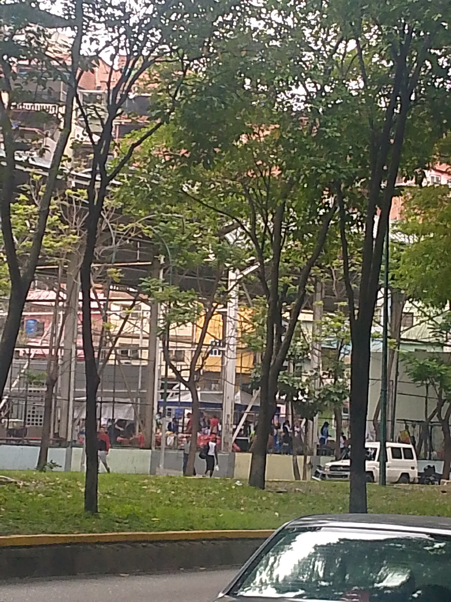 The image shows an outdoor scene in Caracas, Venezuela. In the background is an open-air structure, possibly a sports facility, with people present inside. A row of trees partially obscures the view of the facility. In the foreground, there is a grassy area with a low concrete barrier separating it from the street. A vehicle is visible in the lower portion of the frame. People are walking and standing around the open-air structure. The lighting indicates it is daytime.
