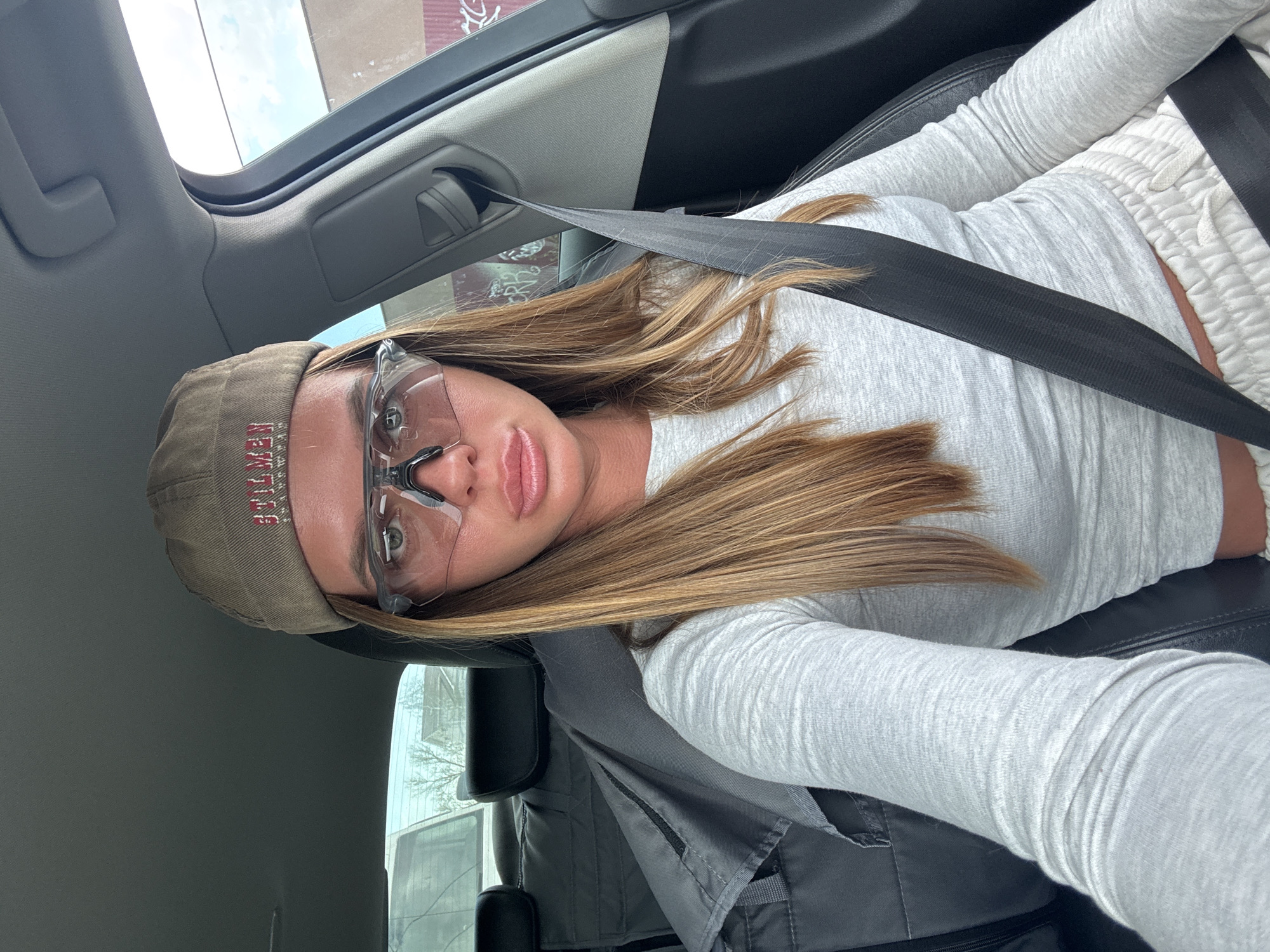 A woman wearing a baseball cap, safety glasses, a light-colored long-sleeved shirt, and light-colored sweatpants is sitting in a car with her seatbelt fastened. The location is identified as Nizhny Novgorod, Russia. The environment includes the interior of a car and a glimpse of the outside through the window.