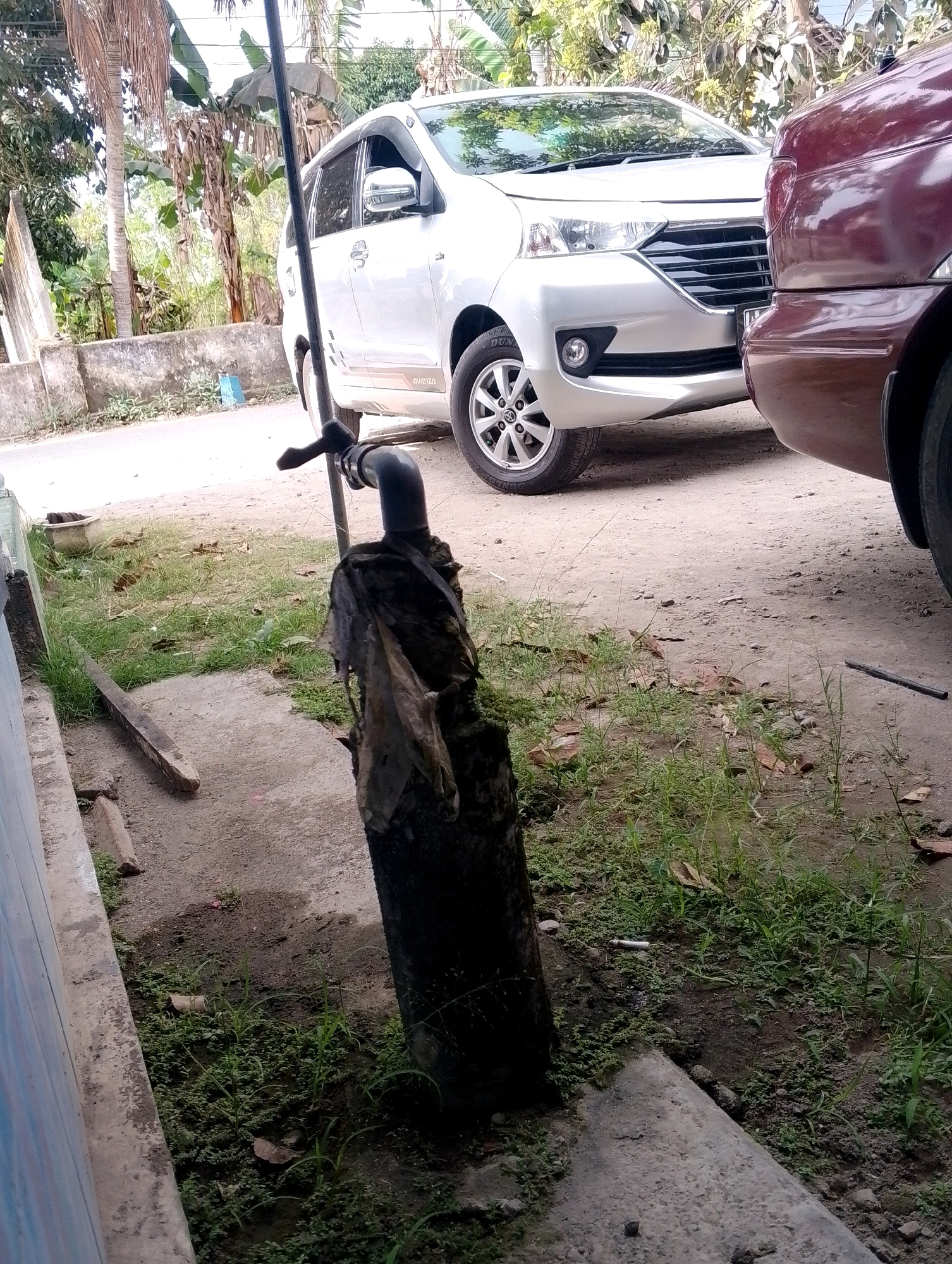 In Ngebra, Indonesia, a street scene unfolds. Two cars, a white Toyota Avanza and a maroon vehicle, are parked on a dirt and concrete surface. In the foreground, a dark, cylindrical object, possibly a pipe or post, is wrapped in dark, tattered material. A thin, black pole extends vertically from the base of this object. The ground is covered with patches of green grass and dry leaves, suggesting a somewhat unkempt outdoor area. In the background, lush green foliage and trees line the street, with a concrete wall visible. The lighting suggests it is daytime.
