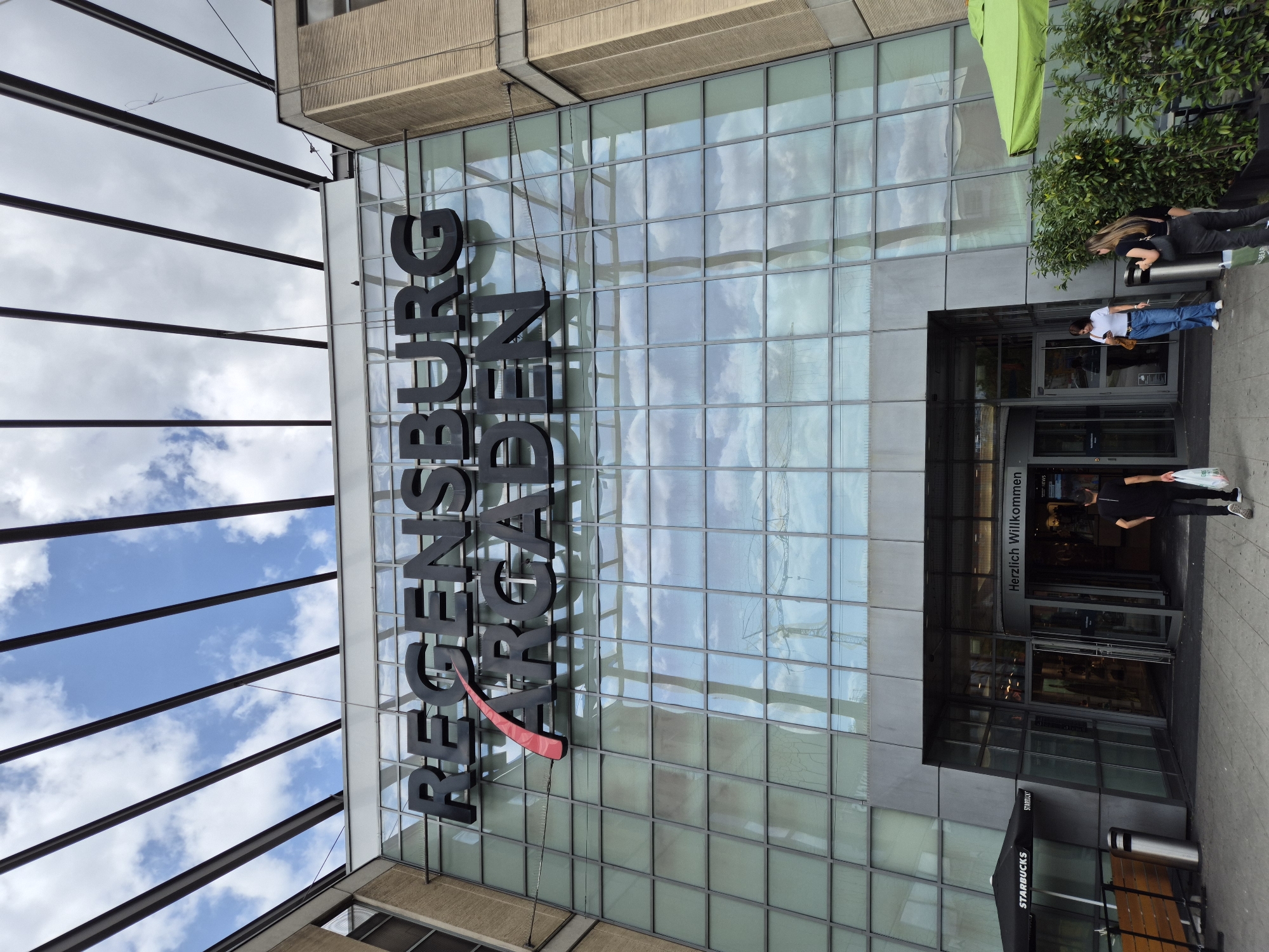 The image shows the Regensburg Arcaden shopping mall in Regensburg, Germany. The mall features a large glass facade with the name "REGENSBURG ARCADEN" prominently displayed in black lettering. Above the facade, a modern metal canopy structure is visible against a cloudy blue sky. At the entrance of the mall, there is a revolving door with a sign above it reading "Herzlich Willkommen" (Welcome). Two people are seen entering the mall. To the left of the entrance, there is a Starbucks outlet with an outdoor seating area. In front of the mall, there are some people and a green umbrella. The overall scene depicts a typical day at a busy shopping center.