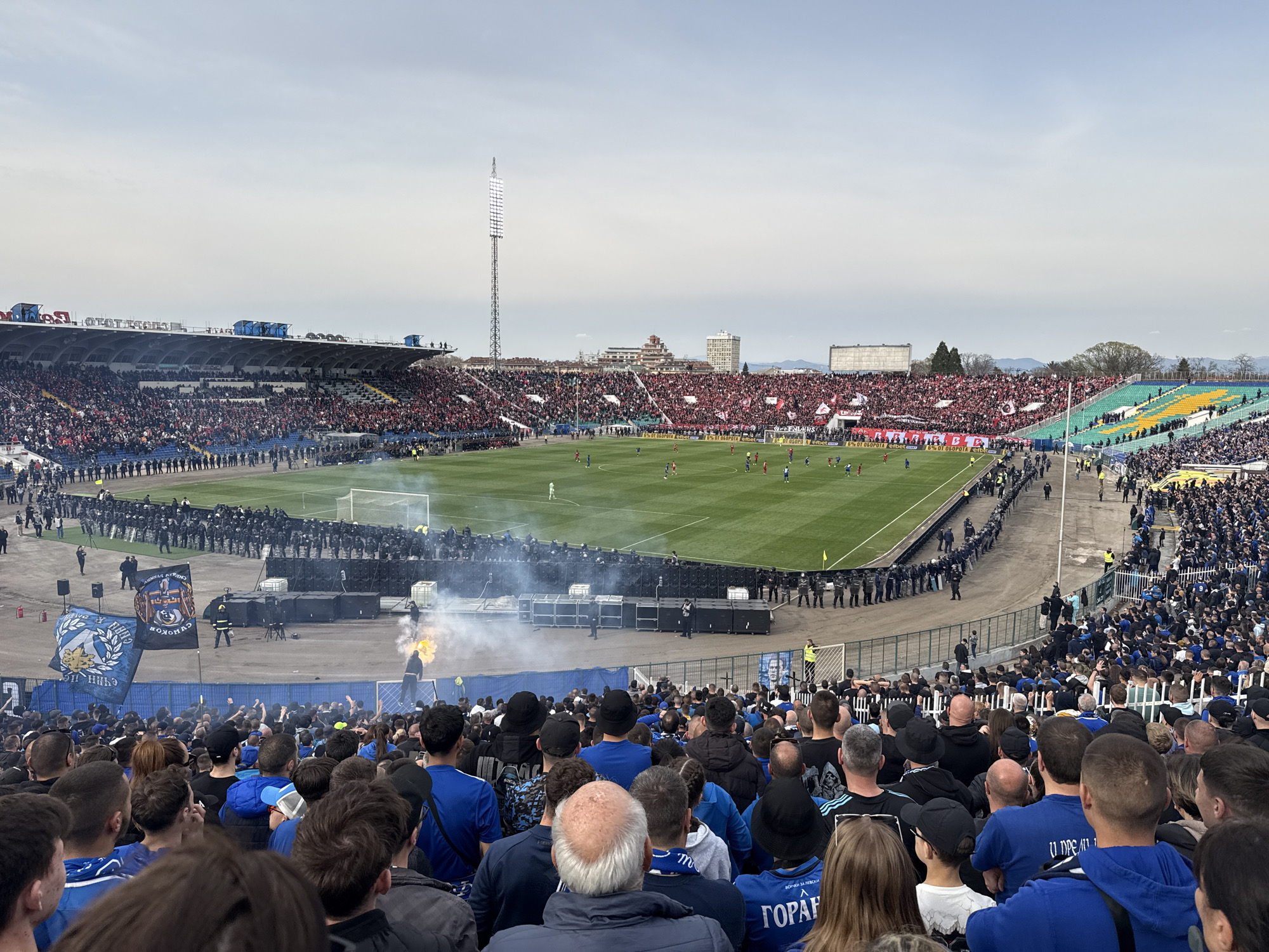 The image shows a soccer match at a stadium packed with spectators. The crowd is segregated into sections, with fans on one side predominantly wearing blue and others wearing red, indicating support for different teams. A line of police in riot gear separates the fans from the field. Smoke from pyrotechnics billows near the field, adding to the atmosphere. The stadium is partially covered with grandstands. In the background, a tall light tower stands beside other buildings. The sky is overcast, casting soft light over the scene. Banners and flags are visible among the crowd, indicating team affiliation.