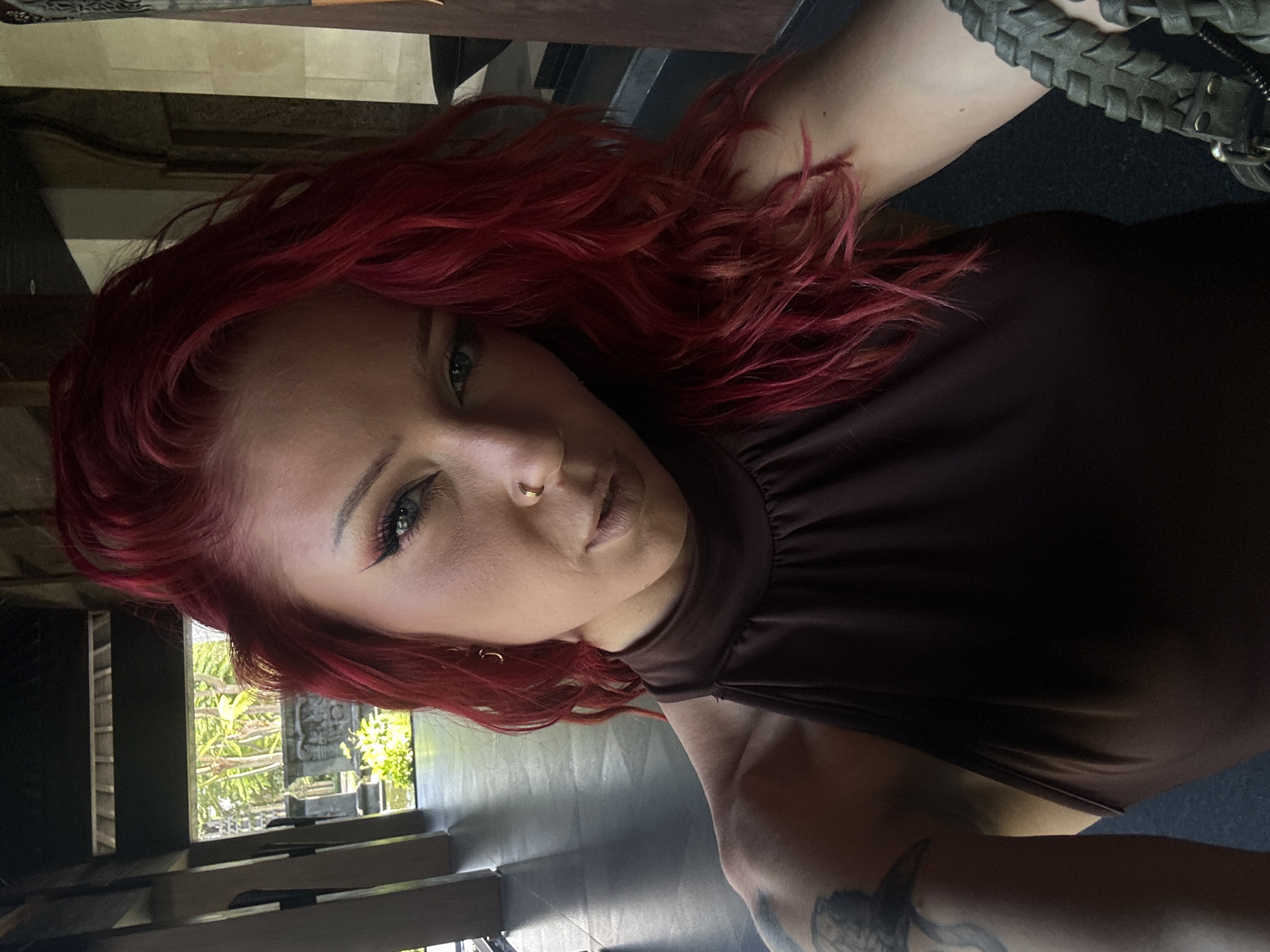 A close-up, angled selfie depicts a woman with vibrant red hair and light eyes. She is wearing a high-necked, dark brown top. The woman's face is partially visible, showing her left eye with winged eyeliner, a nose ring, and light-colored lips. The environment behind her includes a view of a lush, green outdoor scene with stonework, suggesting a tropical garden or temple. The photo's location is indicated as Kuta Selatan, Indonesia. A braided strap, potentially from a bag, is visible in the upper right corner. A partial view of an arm with a tattoo is visible in the lower right of the image.
