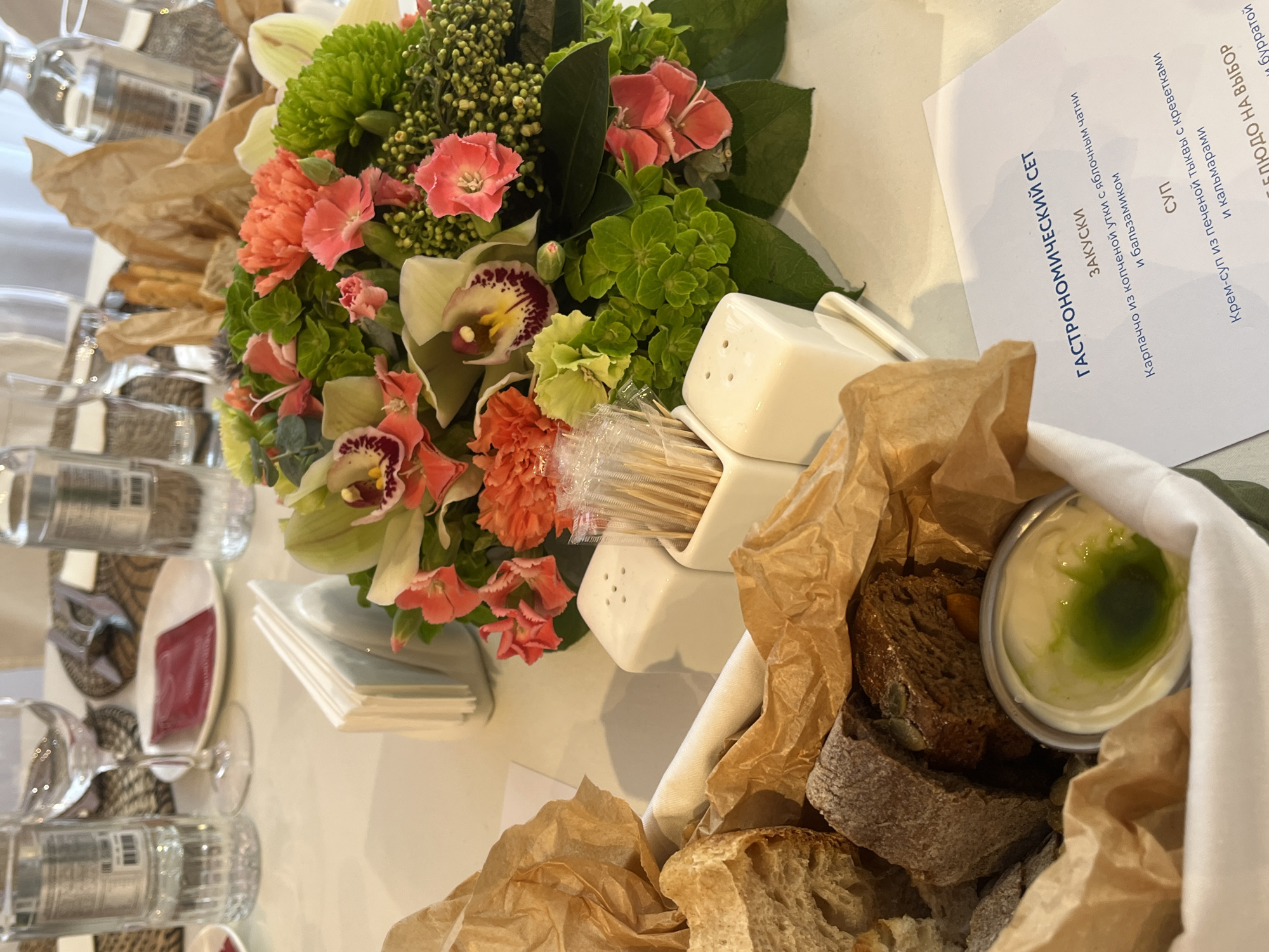 This media file captures a table setting at what appears to be a formal meal or event, likely indoors in Shushary, Russia. The scene is dominated by a vibrant floral centerpiece and a selection of food items.
In the center, a lush floral arrangement features a mix of pink and orange carnations, light green orchids with distinctive purple and yellow markings, and various green foliage including dense clusters of small green buds and larger leaves.
To the right, a white linen-lined basket holds an assortment of bread in brown paper, alongside a small cup of white dipping sauce topped with a green drizzle. Nearby are white square salt and pepper shakers and a container filled with toothpicks.
On the left side of the table, multiple clear glass water bottles with labels and wine glasses are visible, suggesting place settings for several guests. Stacks of white napkins are also placed on the table. The table is covered with a white tablecloth.
A menu card, partially visible and angled towards the viewer, is written in Russian. The visible text details a "ГАСТРОНОМИЧЕСКИЙ СЕТ" (GASTRONOMIC SET) including "ЗАКУСКИ" (APPETIZERS) like "Карпаччо из копченой утки с яблочным чатни и бальзамиком" (Smoked duck carpaccio with apple chutney and balsamic). Under "СУП" (SOUP), "Крем-суп из печеной тыквы с креветками и кальмарами" (Cream soup of baked pumpkin with shrimp and squid) is listed. Further down, "БЛЮДО НА ВЫБОР" (DISH TO CHOOSE) is visible, followed by "бурратой" (burrata), indicating main course options.
The lighting is bright and even, suggesting either daylight or well-illuminated indoor conditions. No people are visible in the frame, but the setup implies a gathering or dining experience.