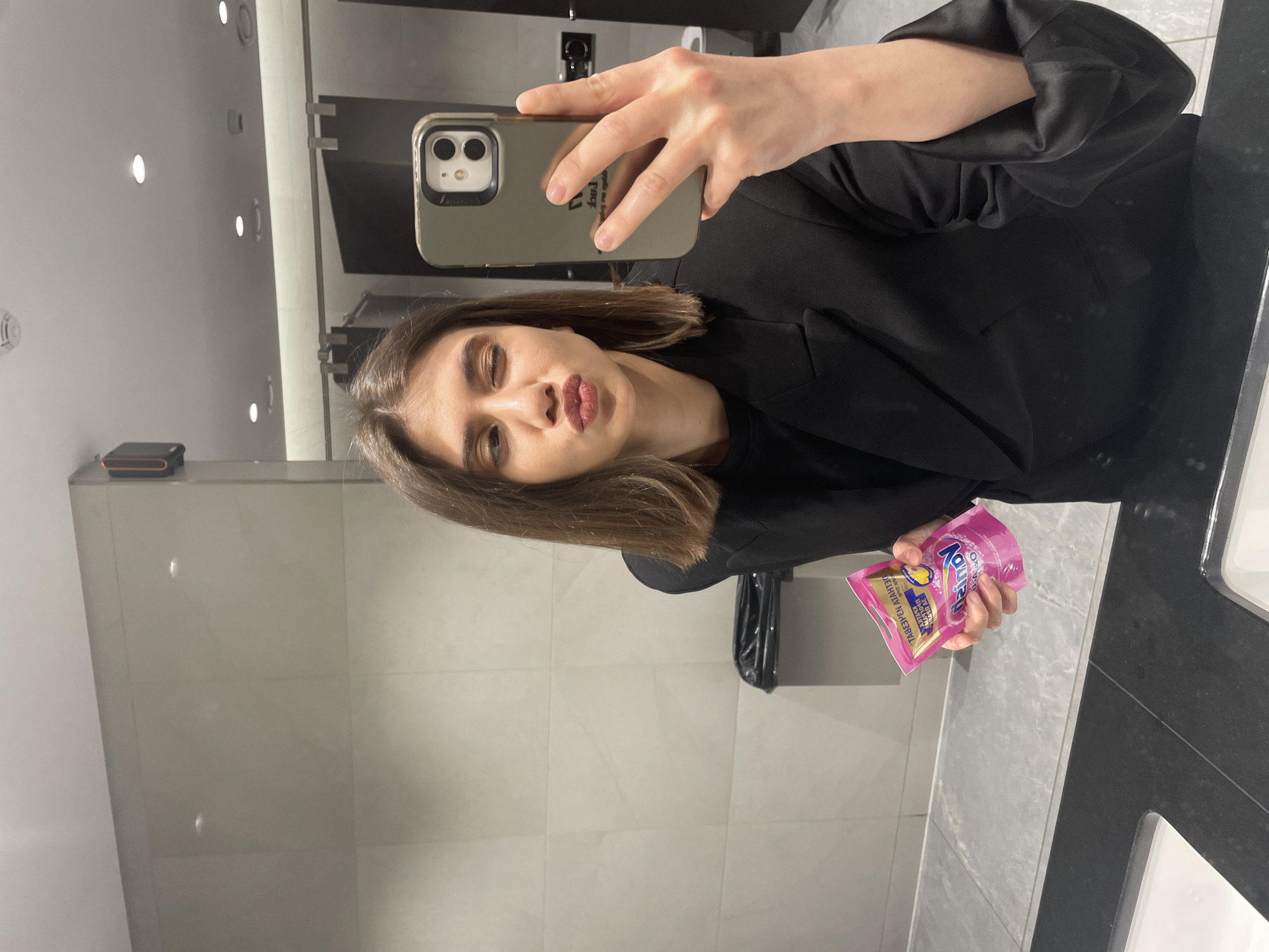 The image shows a woman taking a selfie in a restroom. She is holding a pink packet in her left hand and a smartphone in her right. She is wearing a black jacket. The restroom has light grey tile walls and a black countertop with a white sink. The location is possibly in Sofia, Bulgaria, but this cannot be verified without further information or visual clues.