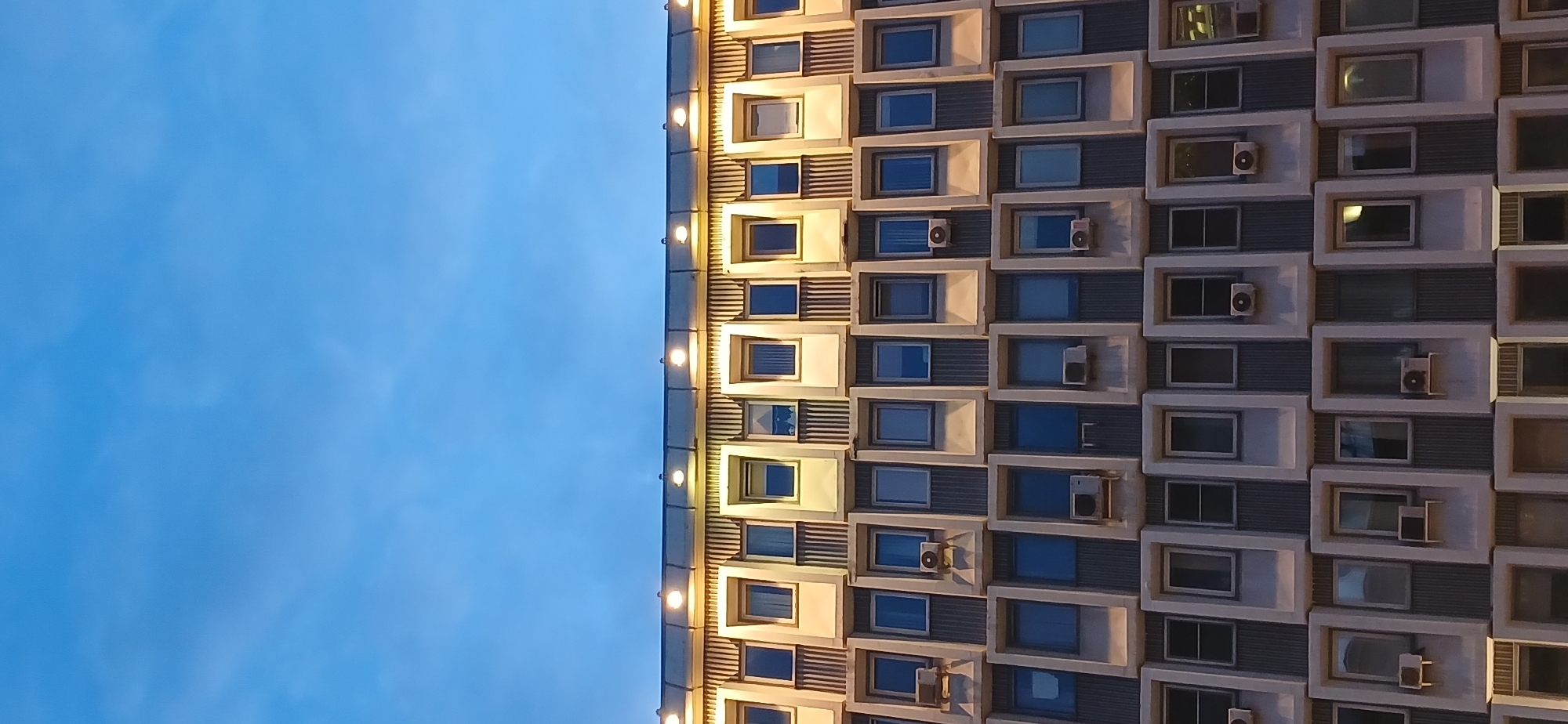 The image shows a tall building with a repetitive pattern of rectangular windows. Many of the windows have air conditioning units installed beneath them. The building is illuminated from above by a row of lights. To the left of the building is a clear blue sky. The building is located in Moscow, Russia.