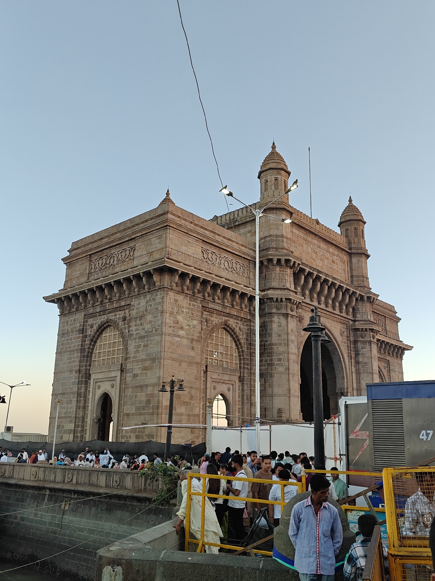 The image depicts the Gateway of India in Mumbai, Maharashtra, India. The monument, made of yellow basalt and reinforced concrete, stands tall against a clear blue sky. A crowd of people gathers around the base of the monument. A section of the area is partitioned by yellow metal railings, beyond which are more people. A dark-colored lamppost stands near the monument. Electrical wires are visible in the sky. Part of an industrial unit is visible on the right.