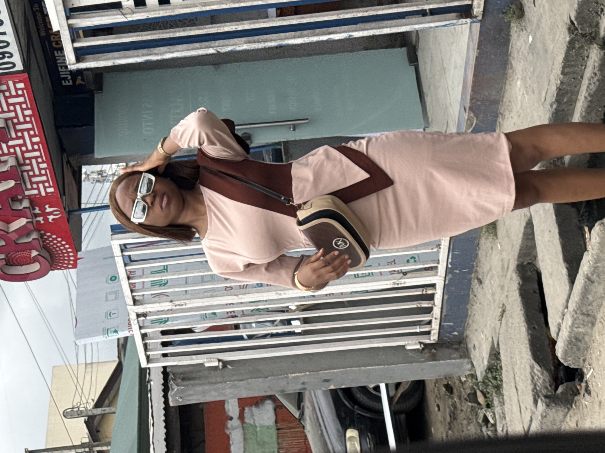 In Ado, Nigeria, a young woman stands on a city street in front of a business establishment with a sign that reads "EJIFINE CRA...". She has dark hair and is wearing a light pink dress with a dark brown overlay. She is also wearing white-rimmed sunglasses and has a crossbody bag with a prominent "MK" logo. Her right hand is touching her head, and her left hand is holding her bag. The street appears to be paved with concrete, and there is some debris and greenery visible on the ground. A black car is parked to the left of the woman, and a metal gate is behind her. The sky is overcast.