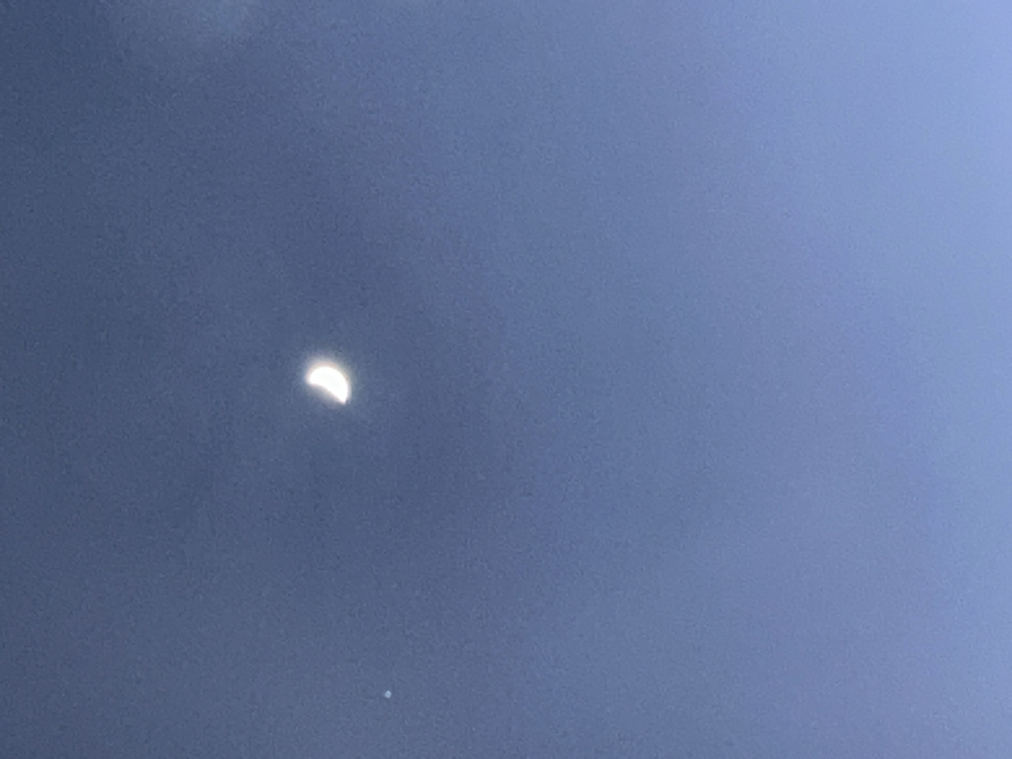 The image captures a clear blue sky over Chaikovsky, Russia, with the moon prominently visible during what appears to be daytime or early twilight. The main subject is a brightly illuminated, thick crescent moon positioned slightly to the left of the center. Its light side faces towards the upper right, indicating the direction of the sun relative to the moon.
Below and slightly to the left of the moon, a small, faint white circular dot is discernible against the blue sky. This could be a bright planet, such as Venus, or a distant star, or potentially a camera artifact.
The sky is a uniform, muted blue, suggesting a clear day with some atmospheric haze, or a moment during sunrise or sunset. There are no visible clouds, land features, man-made structures, or people in the frame. No text is visible. The scene conveys a tranquil observation of a celestial body during daylight hours.