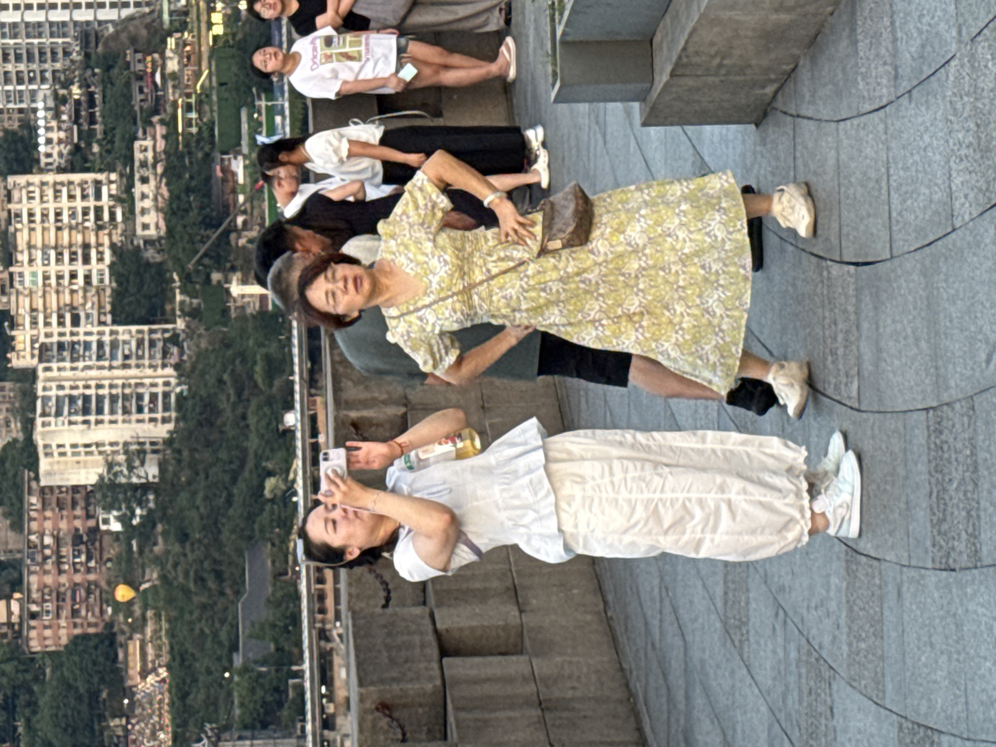 In Yuzhong District, China, two women are standing on a paved walkway with a stone barrier to their left. The woman on the left is wearing a white outfit consisting of a ruffled top and loose pants, and is holding a smartphone, appearing to take a picture. She has a bottle of yellow liquid hanging from her wrist. The woman on the right is wearing a yellow and white floral dress and a small brown crossbody bag. She is posing with her hands on her hips and looking to her left. In the background, a city with numerous buildings built on a hillside is visible, along with a hot air balloon in the sky. Other people are partially visible in the background, some appearing to be tourists. The overall environment suggests an urban outdoor setting with a scenic view.