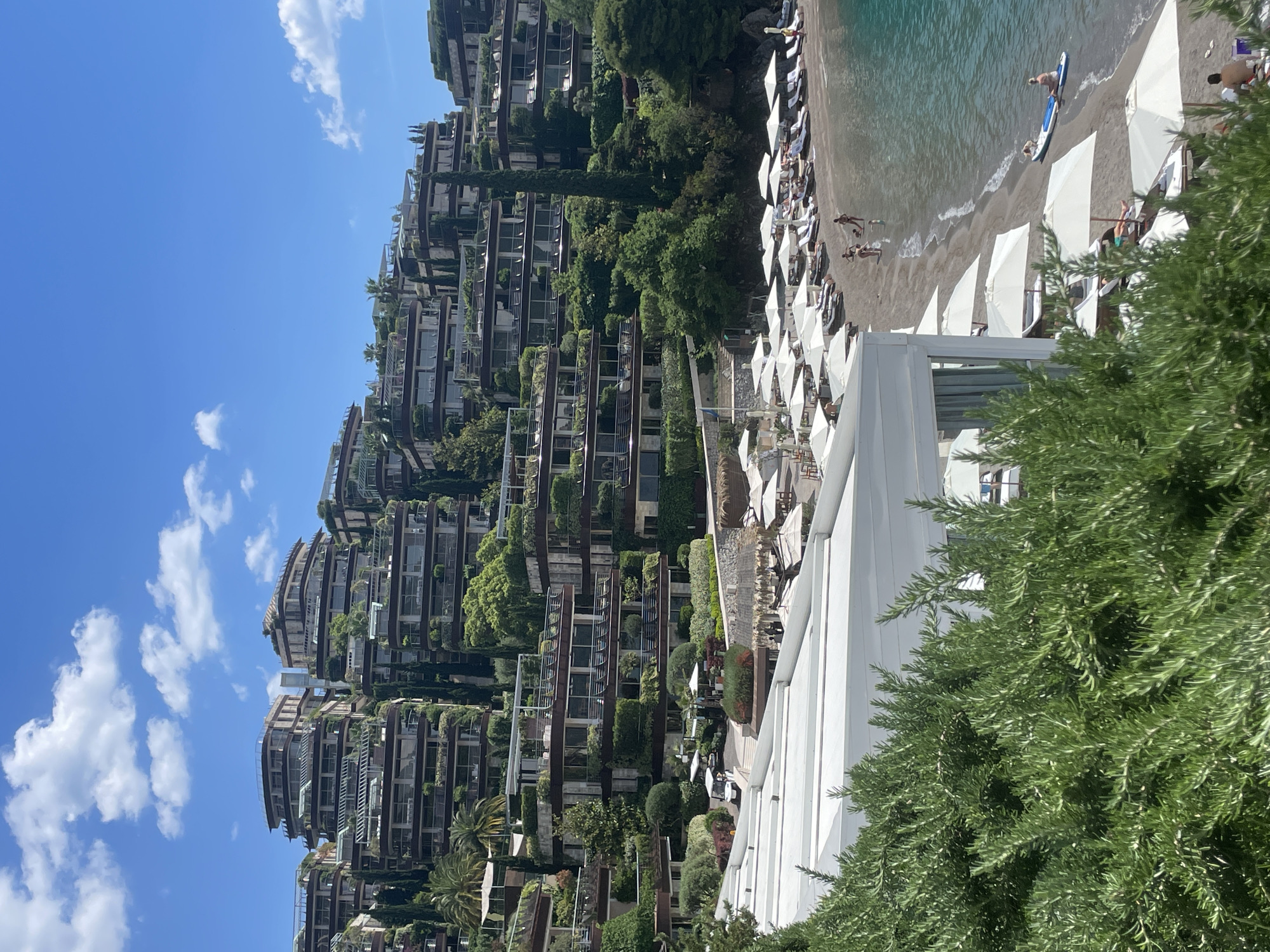 The image depicts a coastal scene in Budva, Montenegro. Tiered residential buildings with green vegetation climb a hillside. A beach is visible with people swimming, sunbathing, and paddleboarding. White umbrellas and lounge chairs populate the beach. The sky is blue with scattered clouds. A large green bush fills the foreground of the image.