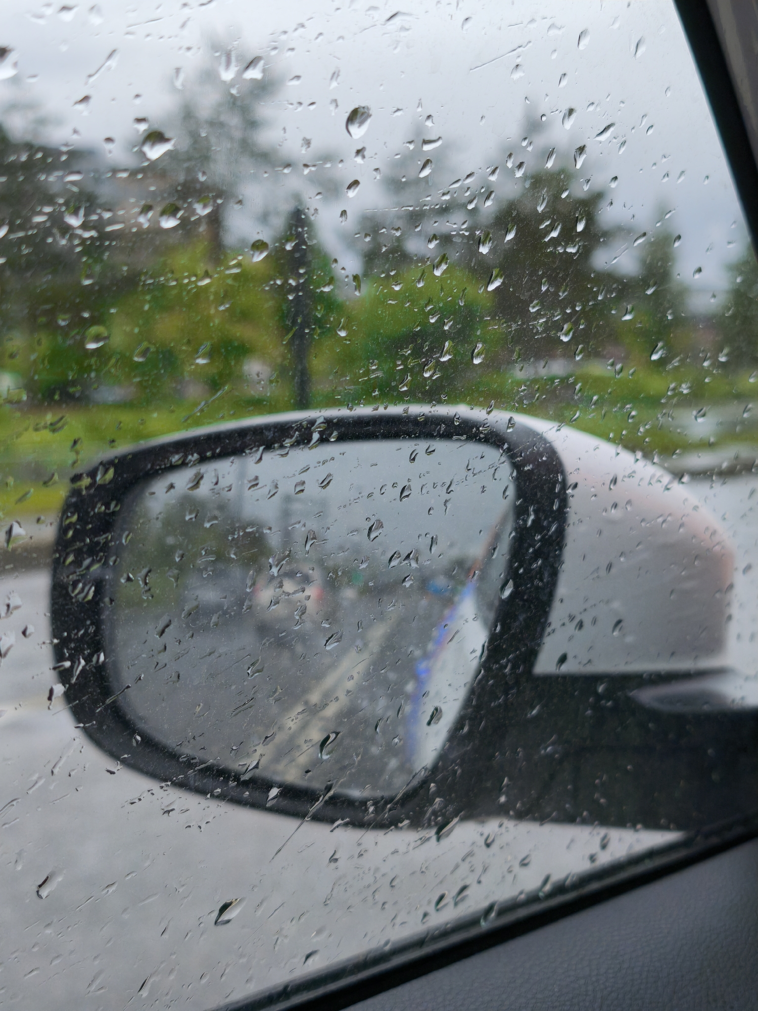The image shows a view through a rain-covered car window. The side mirror is visible, reflecting a wet road and blurred traffic. Outside the window, trees and buildings are visible, also blurred by the rain. No people are visible. The image evokes a rainy day scene. There is no evidence that this image was taken in Moscow, Russia.