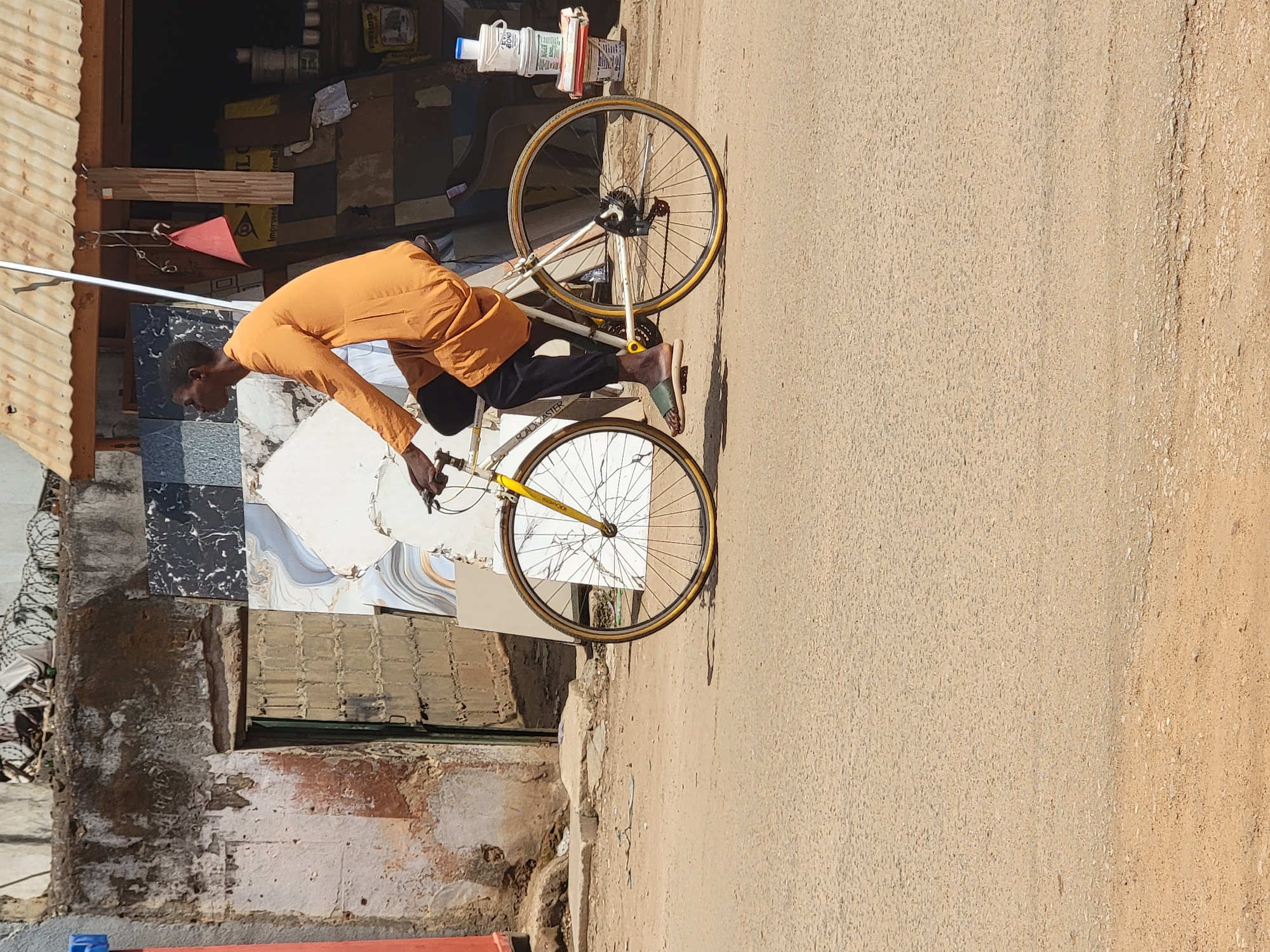 A man wearing an orange long-sleeved shirt and black pants is sitting on a white and yellow bicycle next to a building with a corrugated metal roof. The building appears to be a shop with tiles stacked outside. The location is Maiburiji, Nigeria. The bicycle is on a paved street.