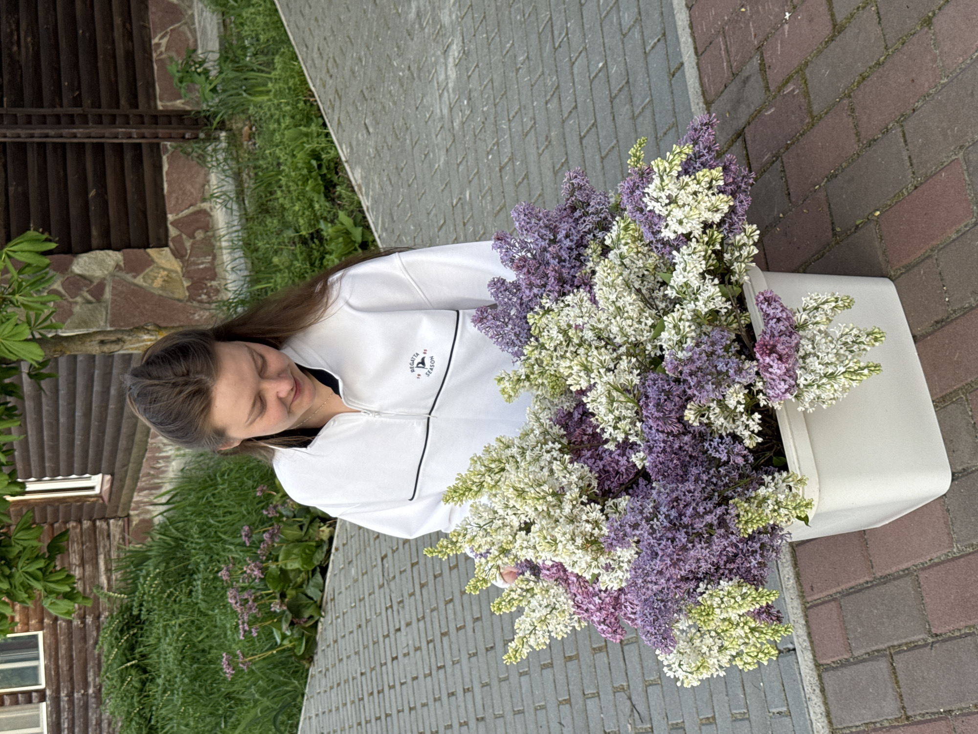A woman is standing outdoors next to a paved area, holding a large white container filled with white and purple flowers, possibly lilacs. The paved area consists of gray bricks on one side and reddish-brown bricks on the other. In the background, there is a building with a wooden facade and green plants. The location appears to be Ленино, Russia.