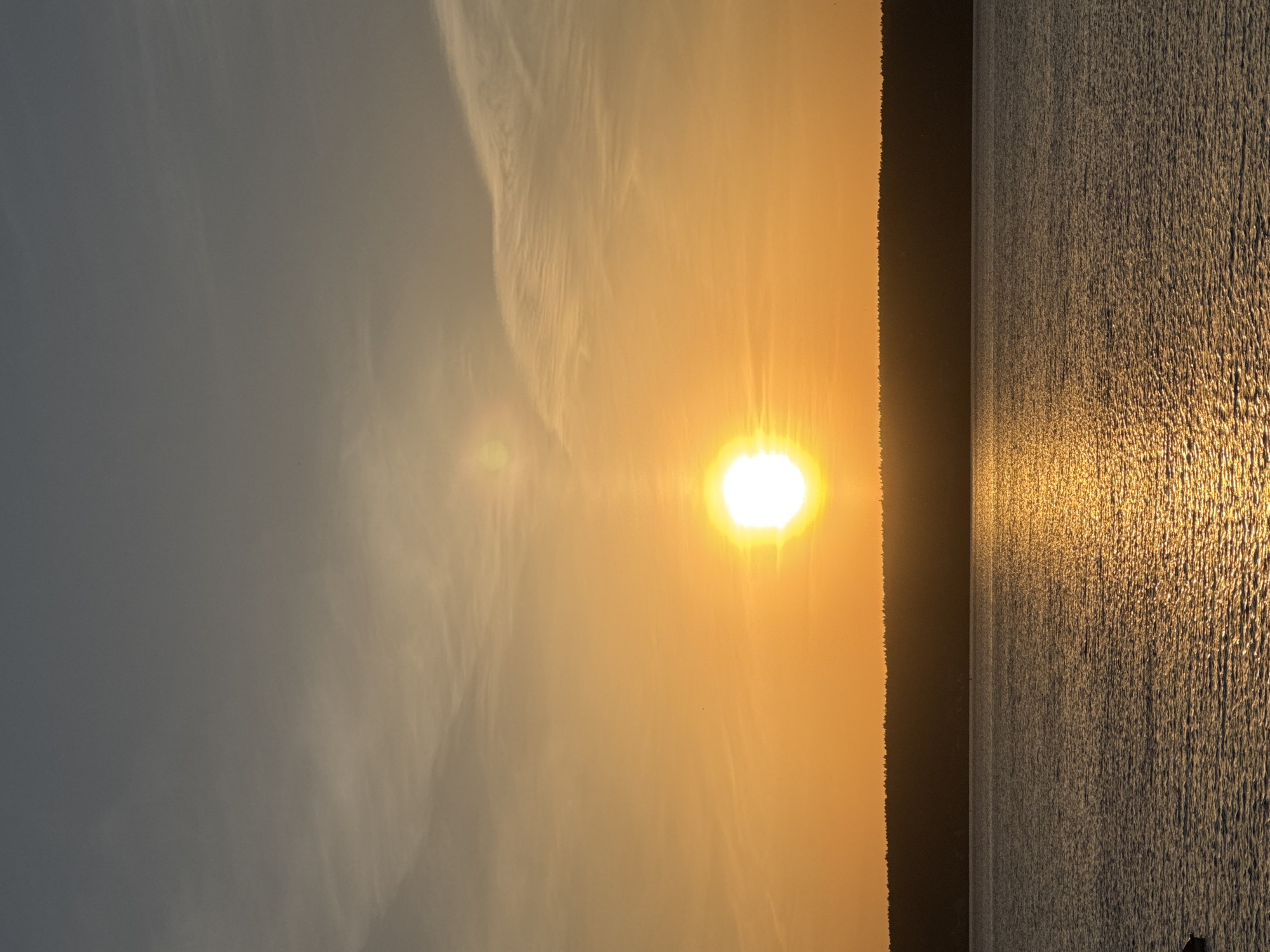 The image depicts a sunset or sunrise scene over a body of water. The sun is positioned in the center of the frame with a distinct golden glow that is reflecting on the water surface, creating a shimmering effect. The sky transitions from a light grey to a warm orange color, indicating the time of day. A dark line, possibly a shoreline or distant land, separates the sky from the water. The exact location is not discernable.