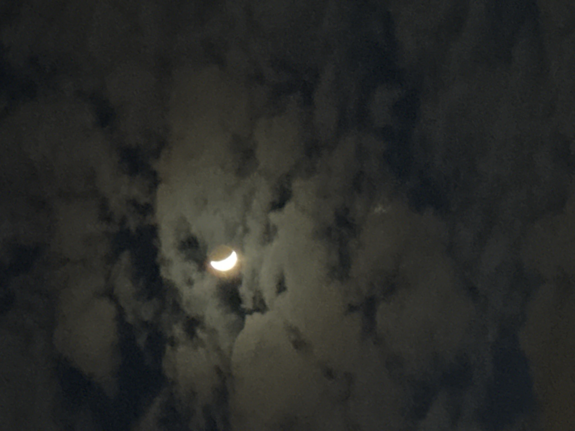 The image shows a crescent moon visible through clouds. There are no people, objects, or actions depicted. The environment is a night sky filled with clouds, obscuring the full moon. The location is not identifiable as Tallinn, Estonia, or any specific city.