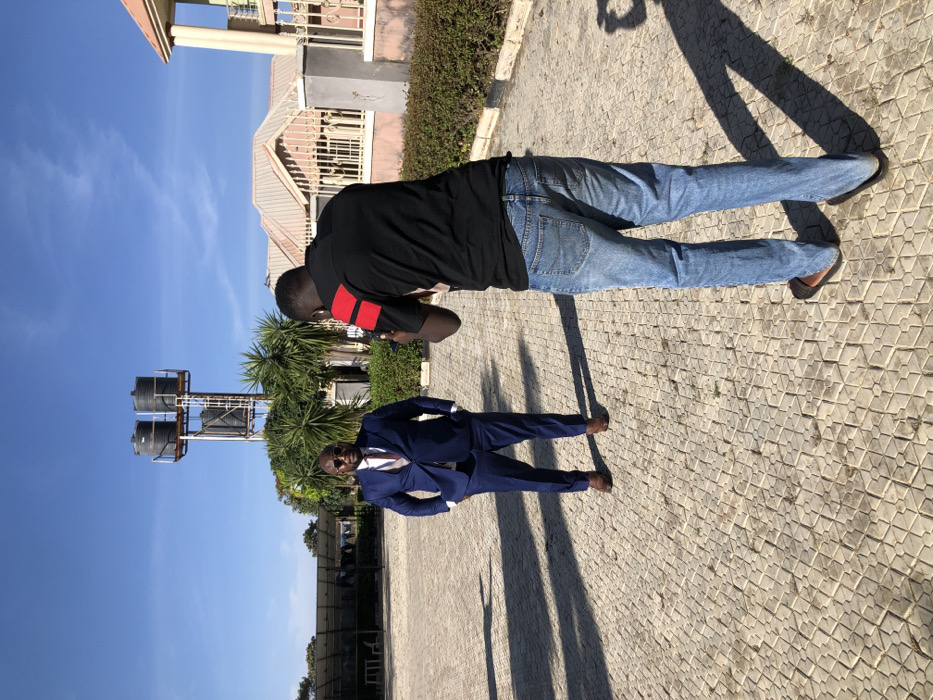Two men stand in a paved courtyard under a clear sky. One, dressed in a blue suit and sunglasses, poses with hands on his hips. Beside him, a second man wears a black shirt with red stripes on the sleeves and blue jeans. In the background are tropical plants, water towers, and a building with a light-colored exterior. The image appears to be rotated 90 degrees counterclockwise. Shadows stretch long from both figures.