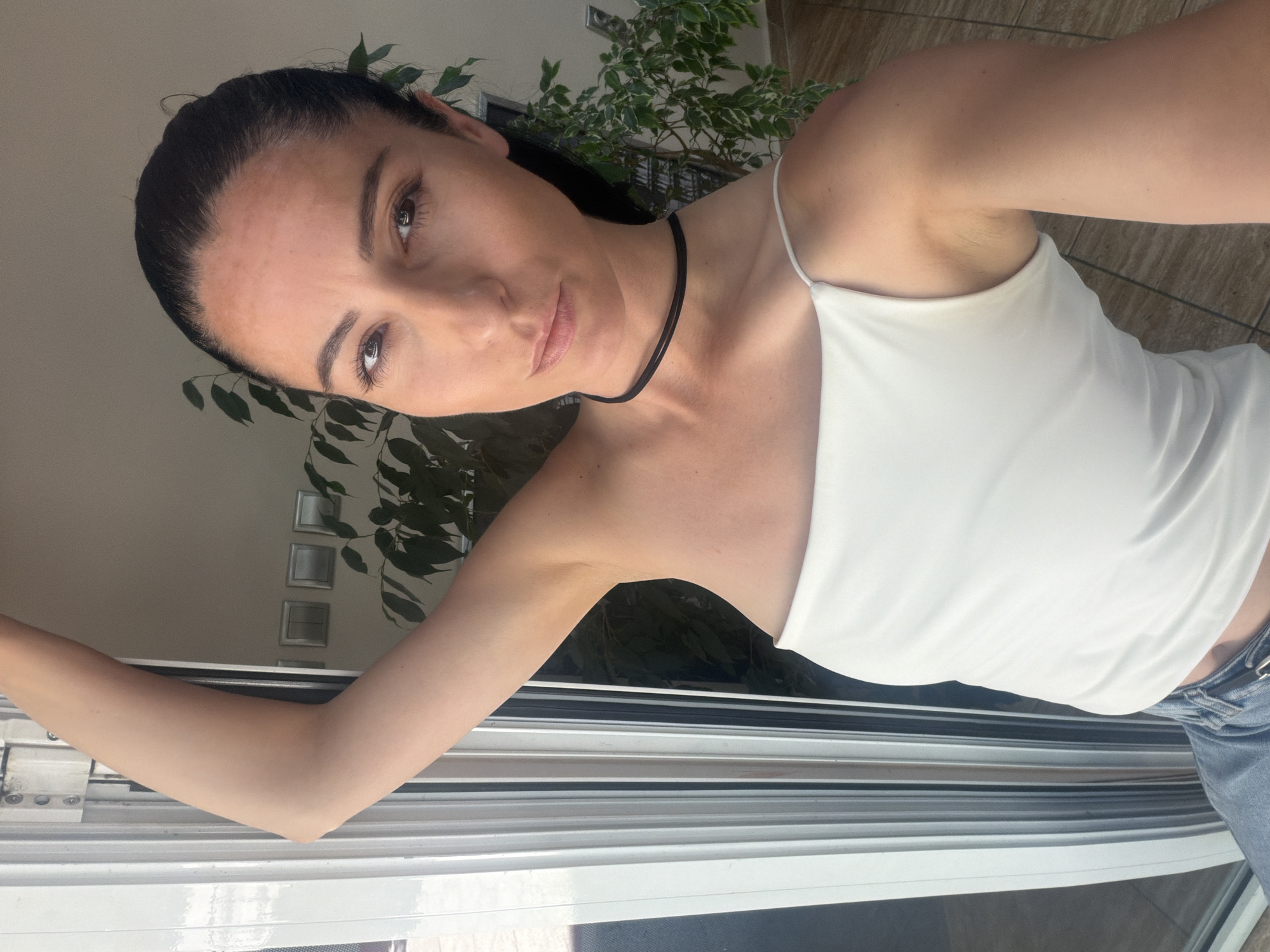 A woman is captured in a selfie taken indoors, possibly in an apartment in Pleven, Bulgaria. She has her left arm raised and bent, with her hand resting on something unseen out of frame. She is wearing a white, spaghetti-strap tank top and blue jeans. A thin black choker necklace is visible around her neck. Her dark hair is pulled back, and she is looking directly at the camera. Behind her, a window with white frames and blinds is partially visible. Inside the room, a potted plant with green leaves is situated to the right of the woman, and a wall with light-colored switches is visible in the background. The lighting suggests it is daytime, with sunlight coming from the window.