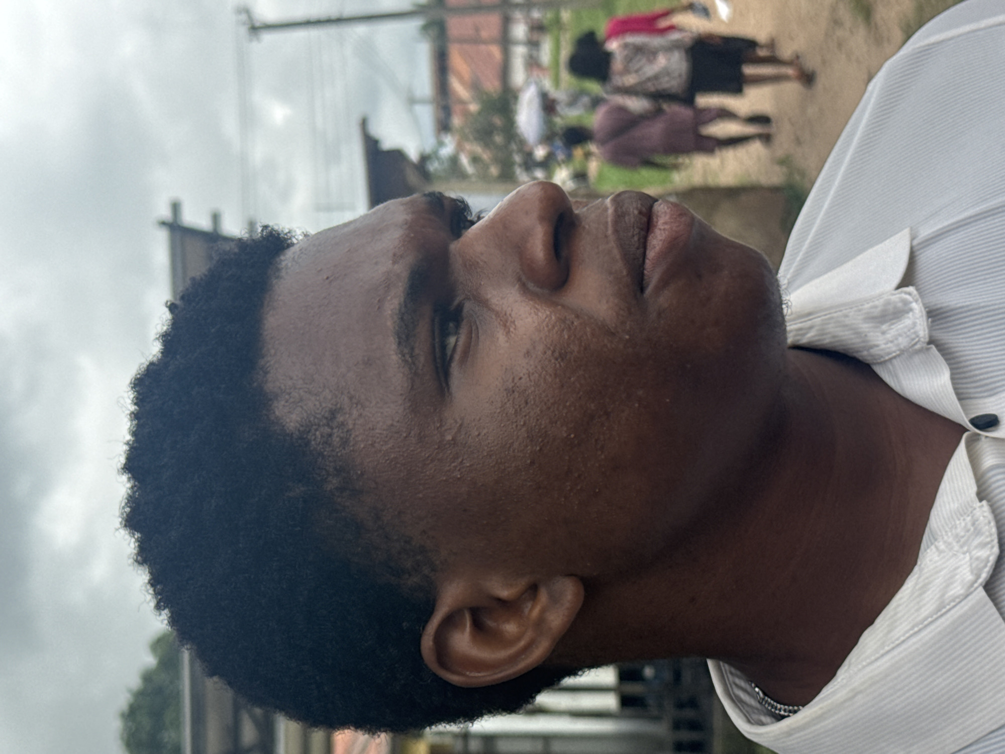 A close-up, profile view captures a young Black man's head and shoulders, angled to his right. He has dark, textured, short-cropped hair and dark skin. His facial features are prominent: his eyes are looking to his right, his nose is broad, and his lips are full. The skin on his face appears slightly oily and has minor blemishes. He is wearing a white collared shirt with thin pinstripes and a silver chain necklace visible at the neckline.
The background is blurred but suggests an outdoor environment with a cloudy sky. Hints of buildings with orange roofs and utility poles are visible. In the mid-ground, out of focus, are several figures walking away from the camera, suggesting a public space. One figure appears to be holding a white umbrella. The ground appears to be a dirt path. The scene is likely located in a town or village setting.