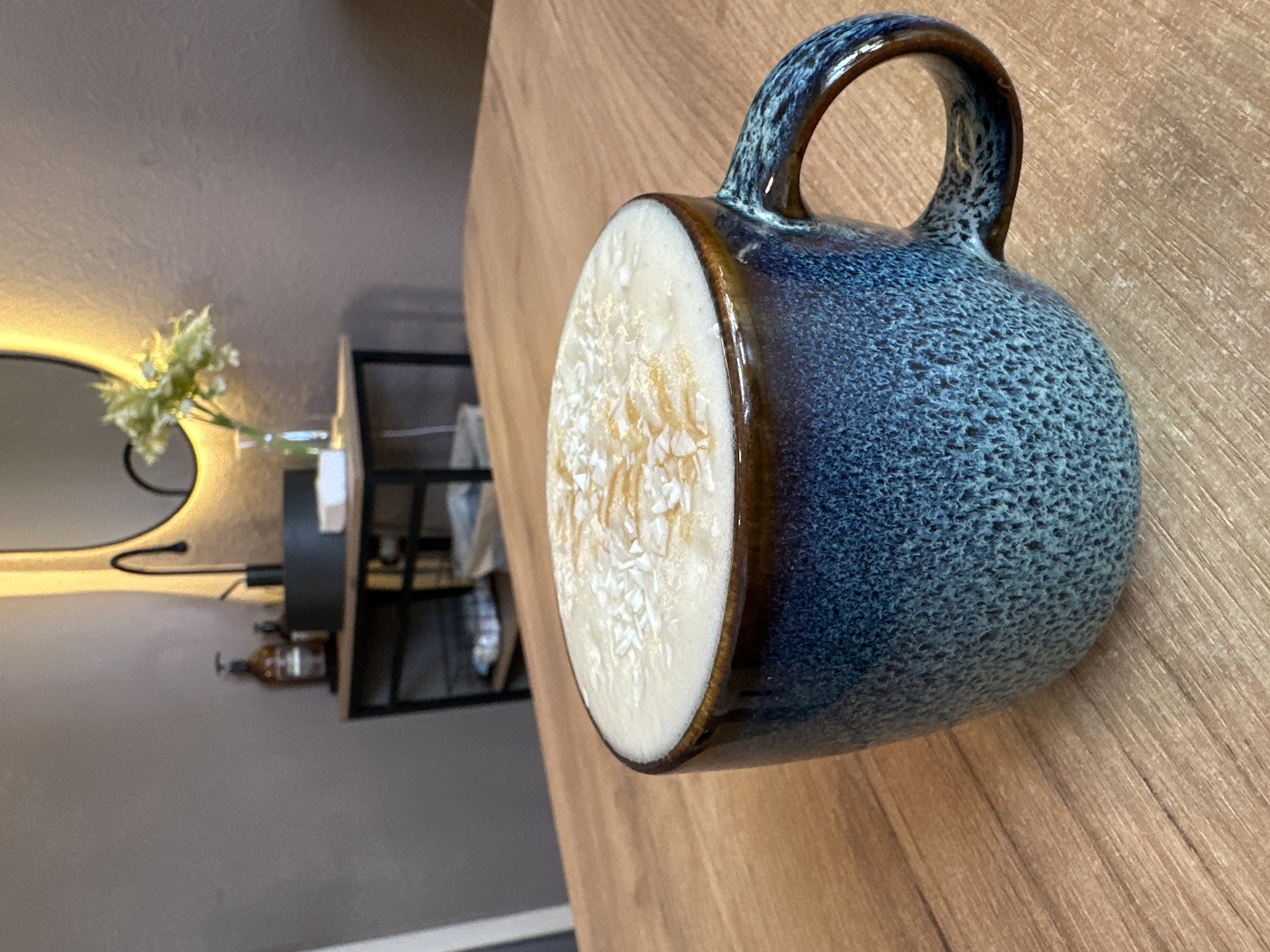 A close-up shot of a blue, speckled ceramic mug, filled with a white liquid topped with a light brown pattern, sits on a light brown wood-grain surface. The mug has a distinctive handle with a dark brown rim around its opening. In the background, out of focus, is a dimly lit area with a round mirror surrounded by a light, a vase with white flowers, a small shelf with various items, and a bottle. The location cannot be determined from the image.