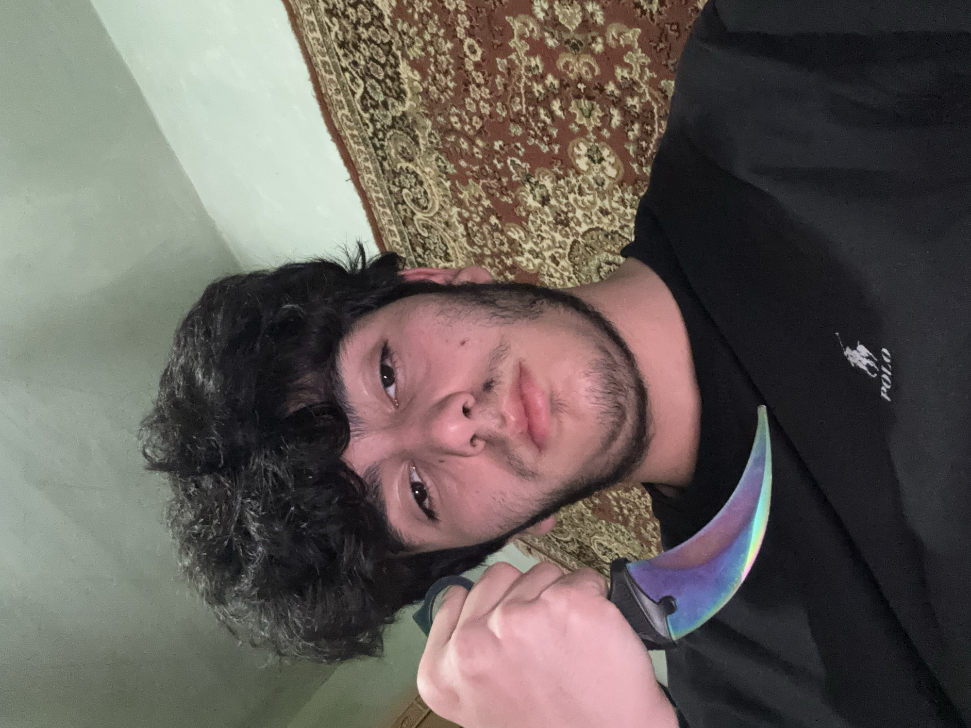 A person is holding a curved iridescent knife. The person has dark curly hair, dark eyes, and a beard, and is wearing a black polo shirt. A rug is visible in the background. The setting appears to be indoors, near a wall. The location and city cannot be determined from the visual data.