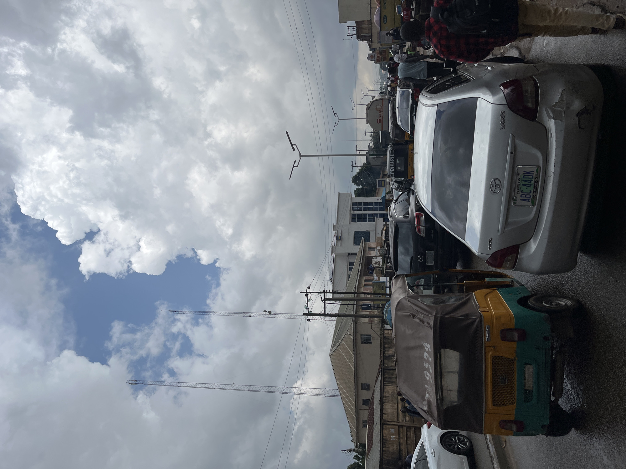 The image depicts a street scene in Jos, Nigeria, characterized by heavy traffic and a cloudy sky. In the foreground, a three-wheeled auto-rickshaw, painted yellow and green with a brown canopy, is stopped in traffic. To its right, a silver sedan with a Nigerian license plate reading "ABUJA ABC 440K" is also stationary. A man in a red and black checkered shirt and beige pants walks past the sedan, carrying a backpack. Further ahead, other vehicles, including a white sedan, a black SUV, and a tanker truck, are visible amidst the congestion. In the background, buildings line the street, and two tall transmission towers stand prominently against the sky. Power lines crisscross the scene, and streetlights are mounted on poles. The ground appears wet, suggesting recent rain or damp conditions. The overall atmosphere is one of urban bustle and movement, typical of a busy commercial area.